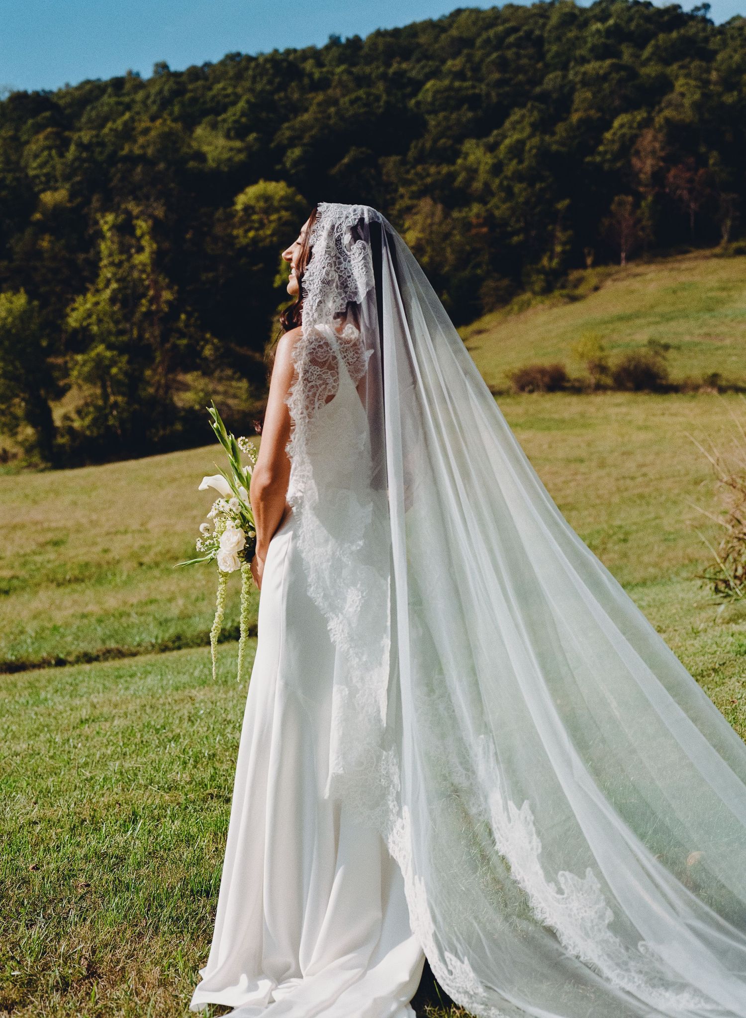 A bride in a lace wedding gown with flowing cathedral veil stands in a scenic mountain meadow on her wedding day.