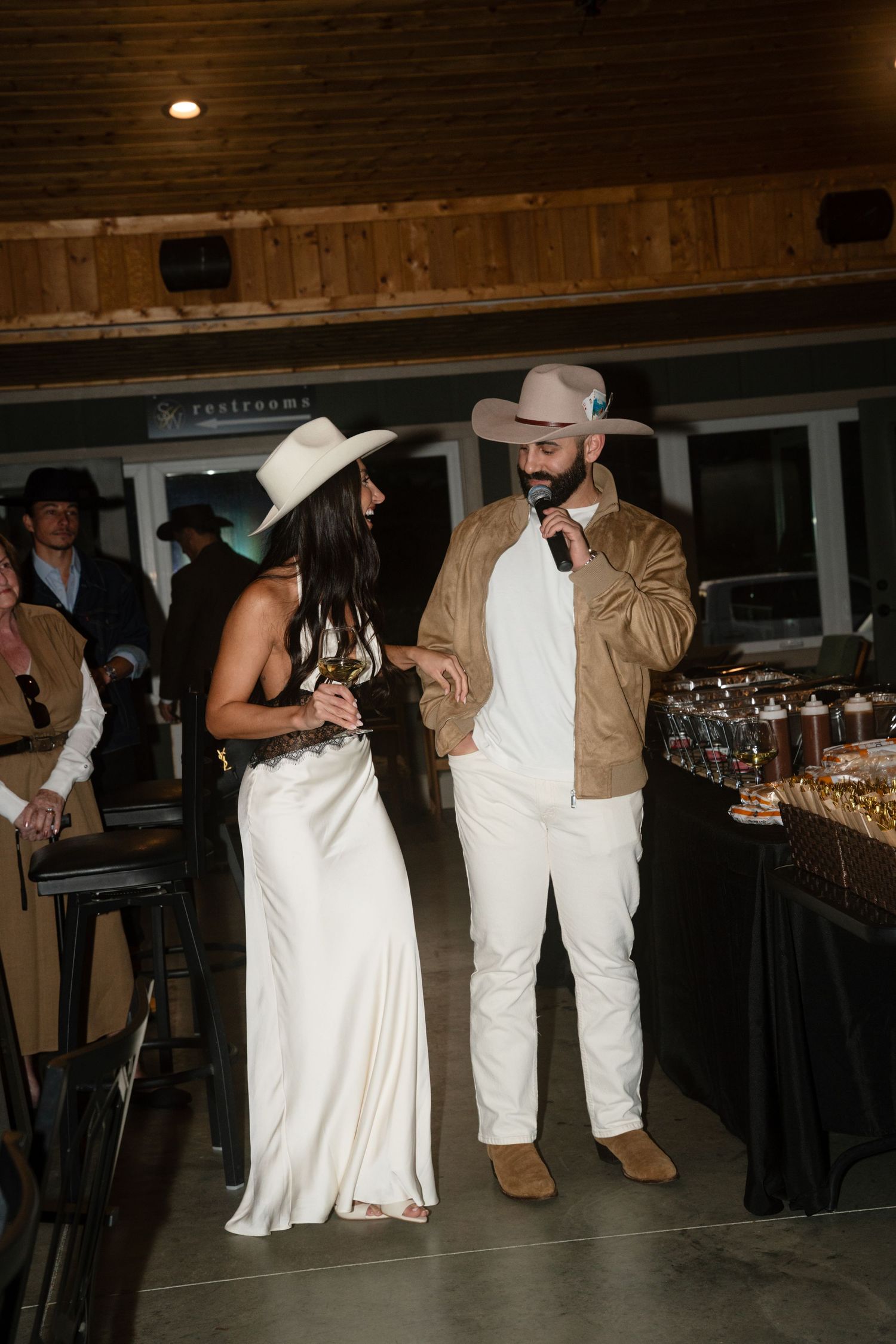 Couple in white outfits and cowboy hats share a mic at a western-themed wedding reception indoors.