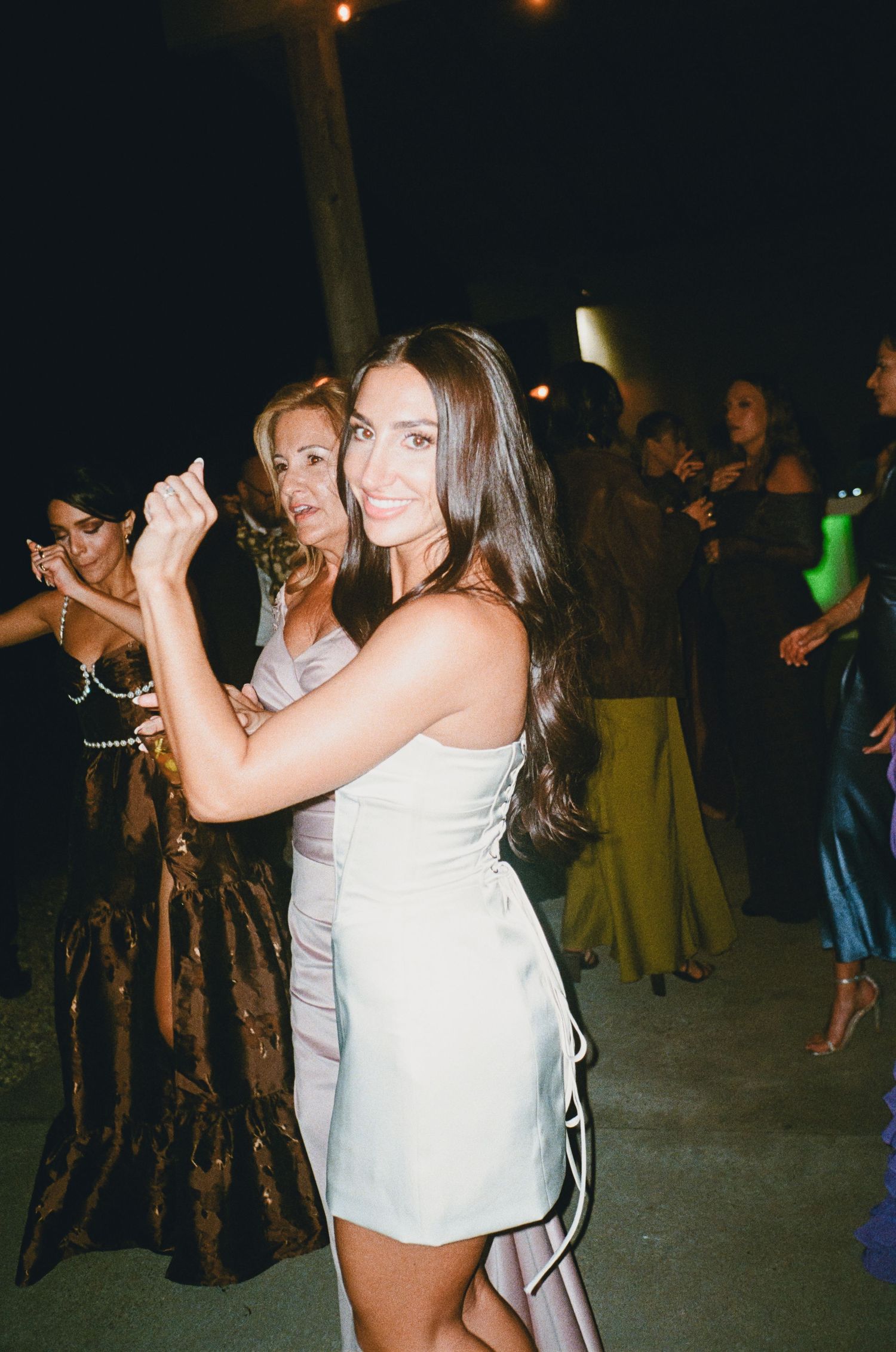 A smiling brunette woman in a white mini dress poses at an elegant outdoor evening party with guests dancing behind her.