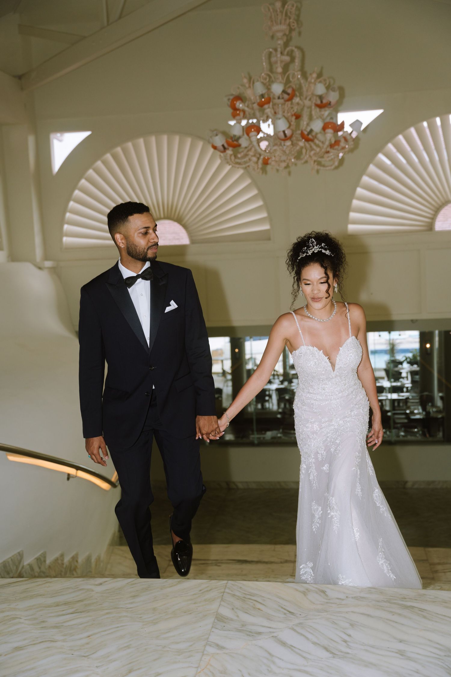 Elegant couple in formal wedding attire pose together in a bright indoor venue with decorative wall fans.