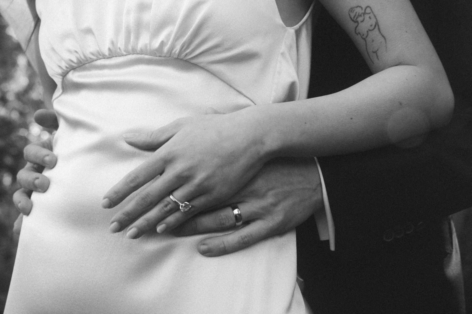 Black and white photo of a pregnant bride and groom embracing, showing wedding rings and a floral tattoo on the groom's arm.