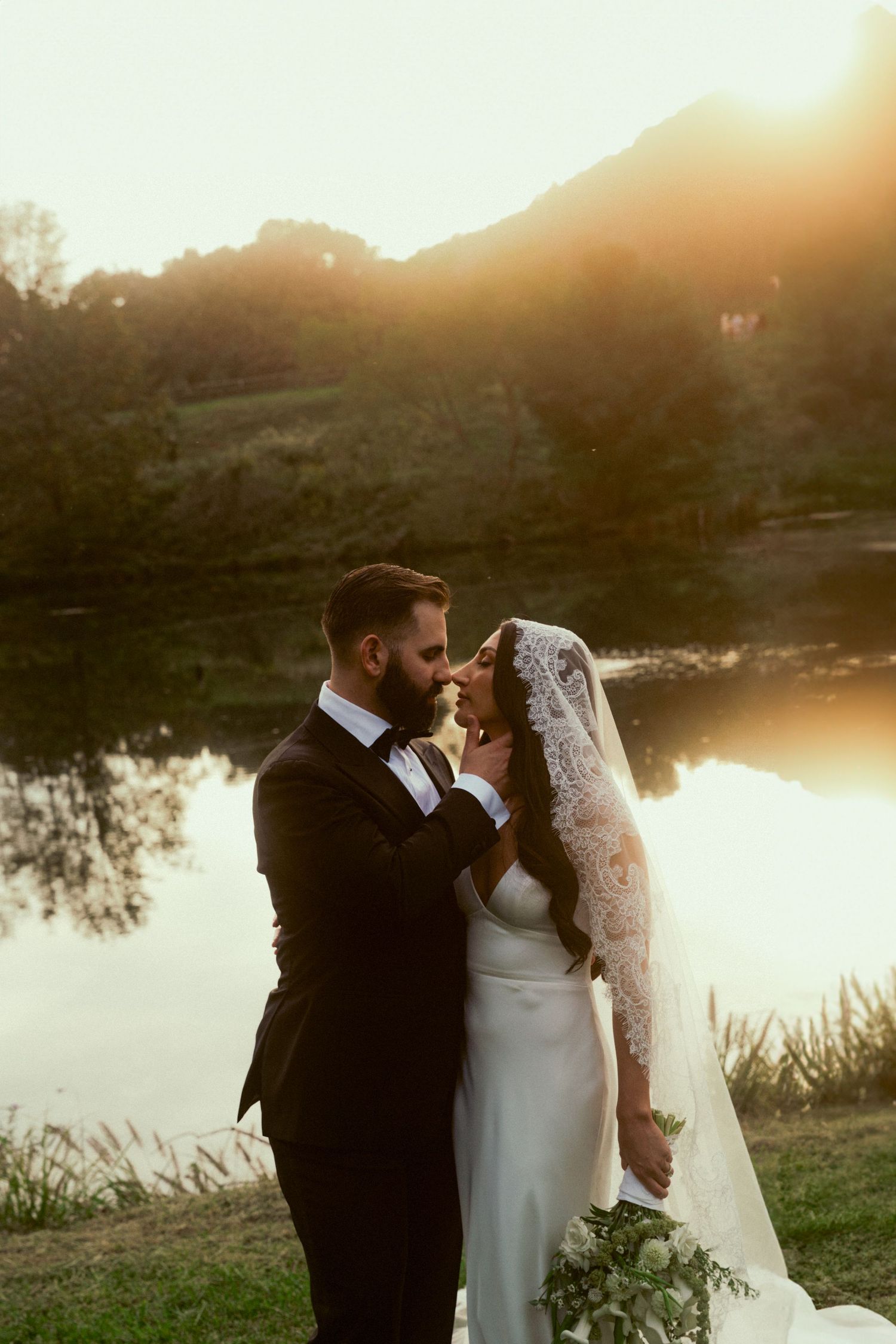 Bride and groom share a romantic moment by a lake at golden sunset, with mountains in the background.