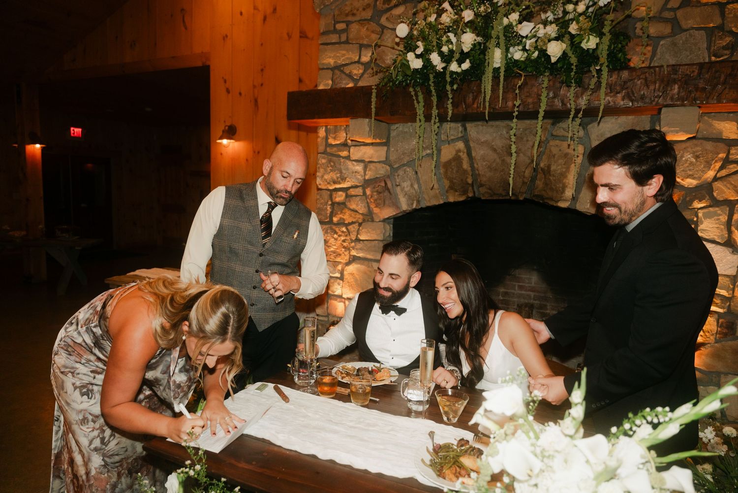 Wedding guests gather around couple signing marriage documents at rustic stone fireplace venue.