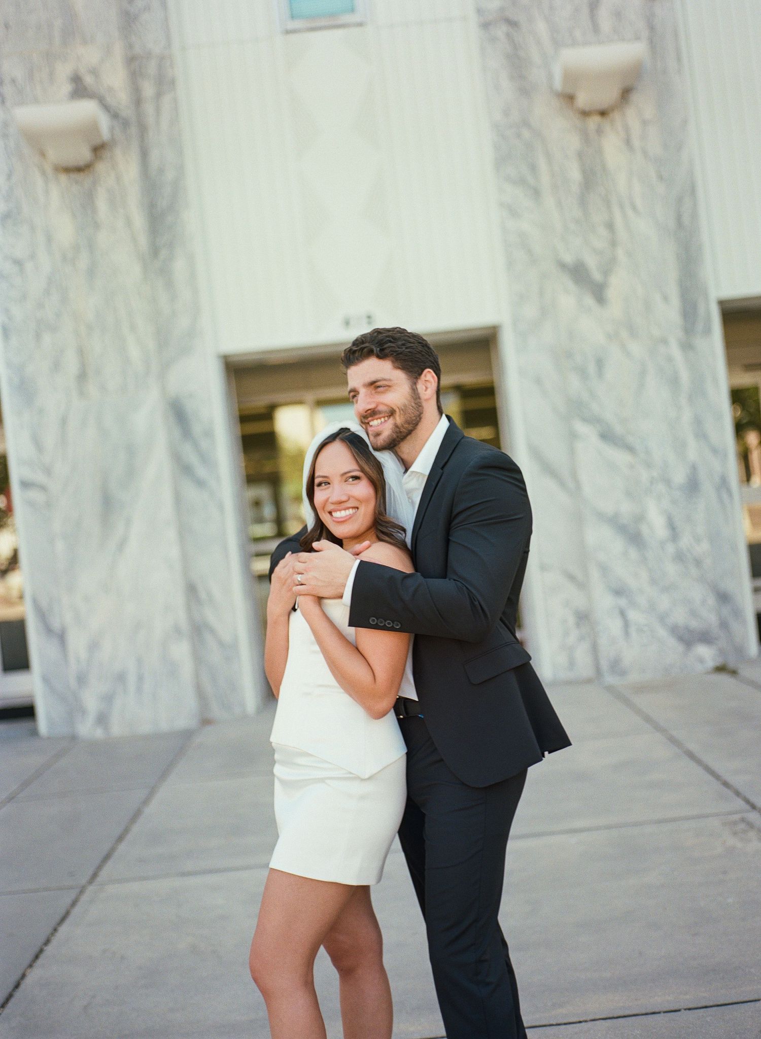 A couple shares a romantic moment in formal attire against a marble wall backdrop.