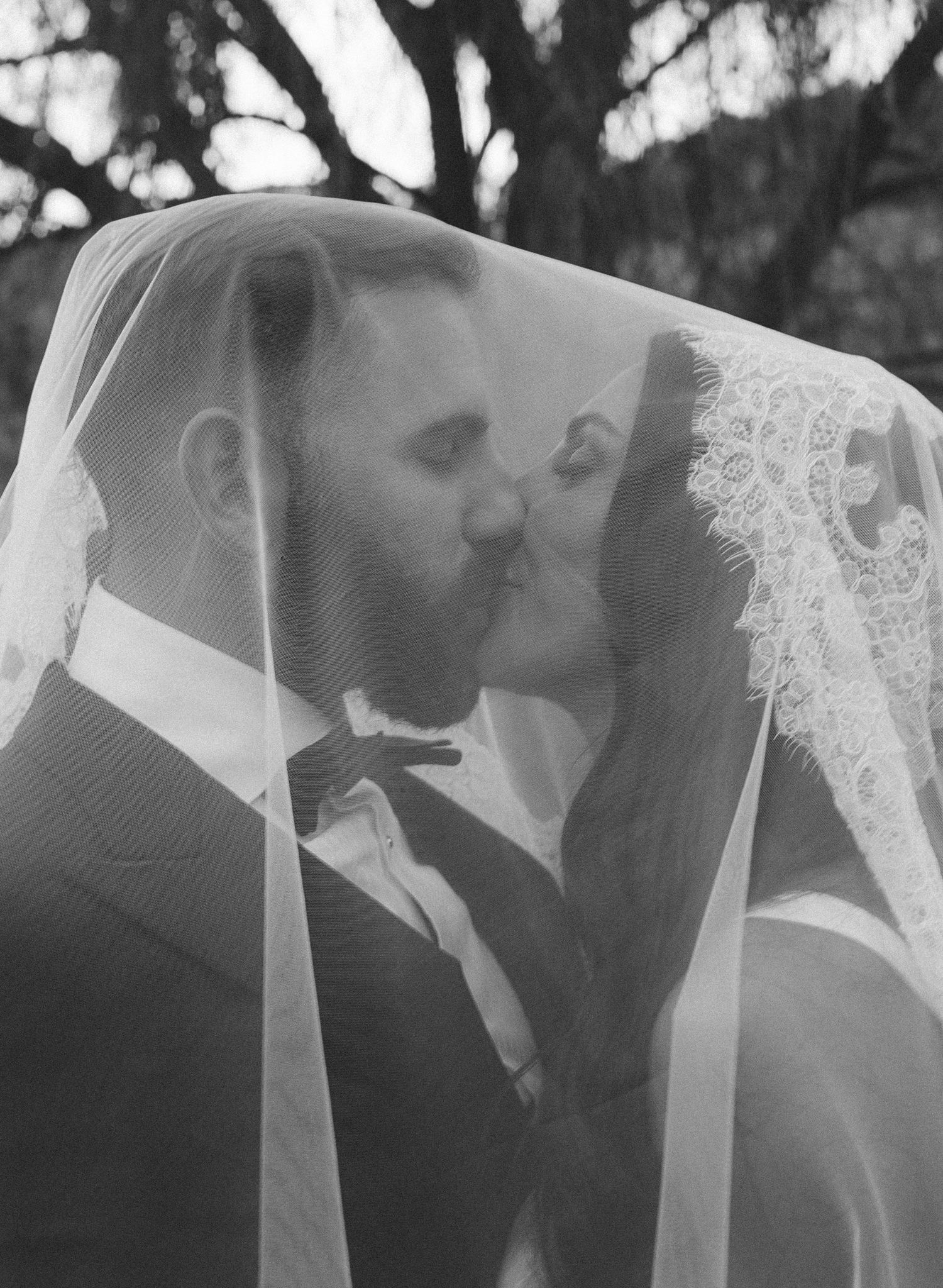 Black and white photo of bride and groom sharing a kiss under a lace veil outdoors.