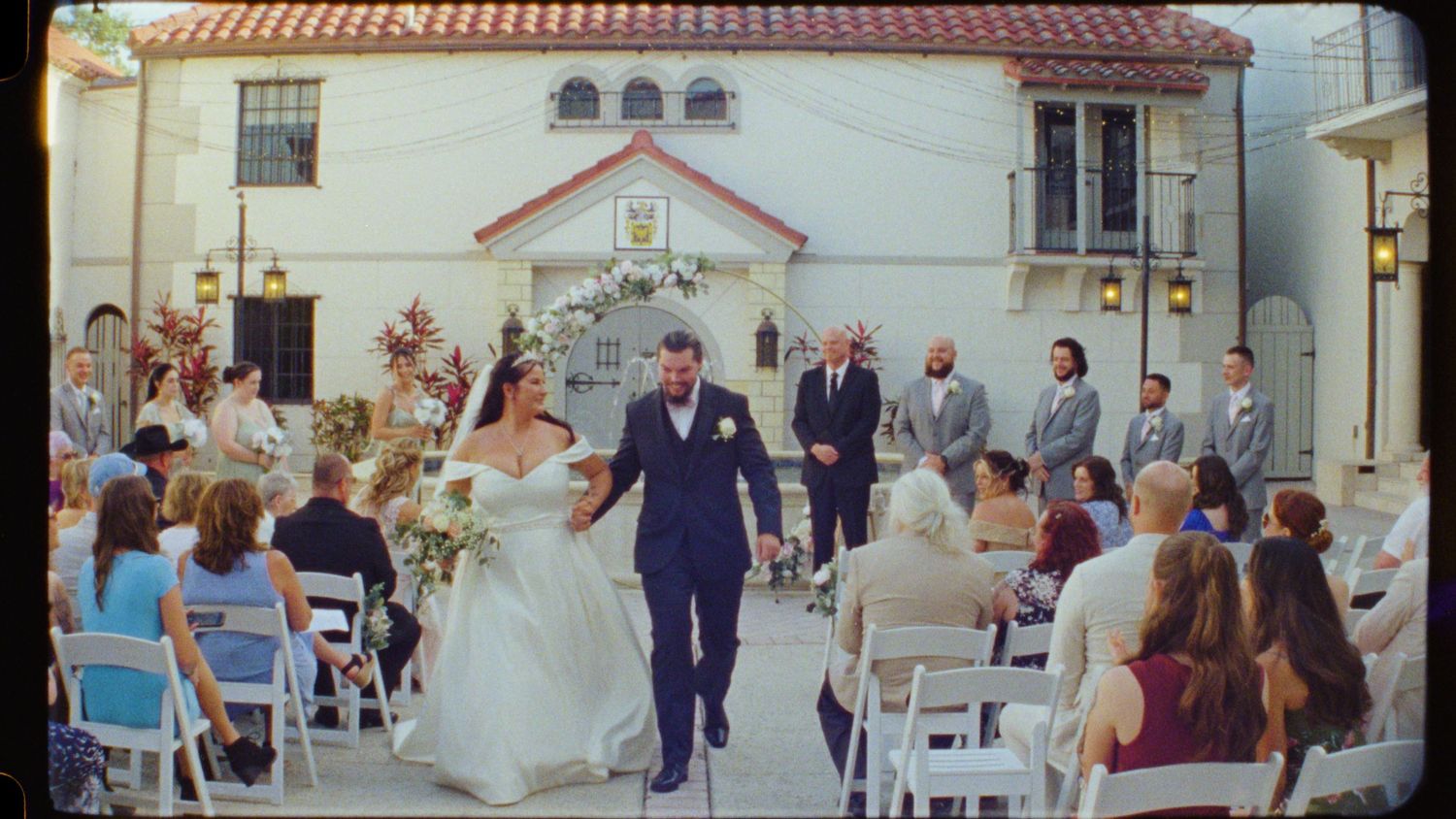 An outdoor wedding reception with white chairs and Spanish-style architecture.