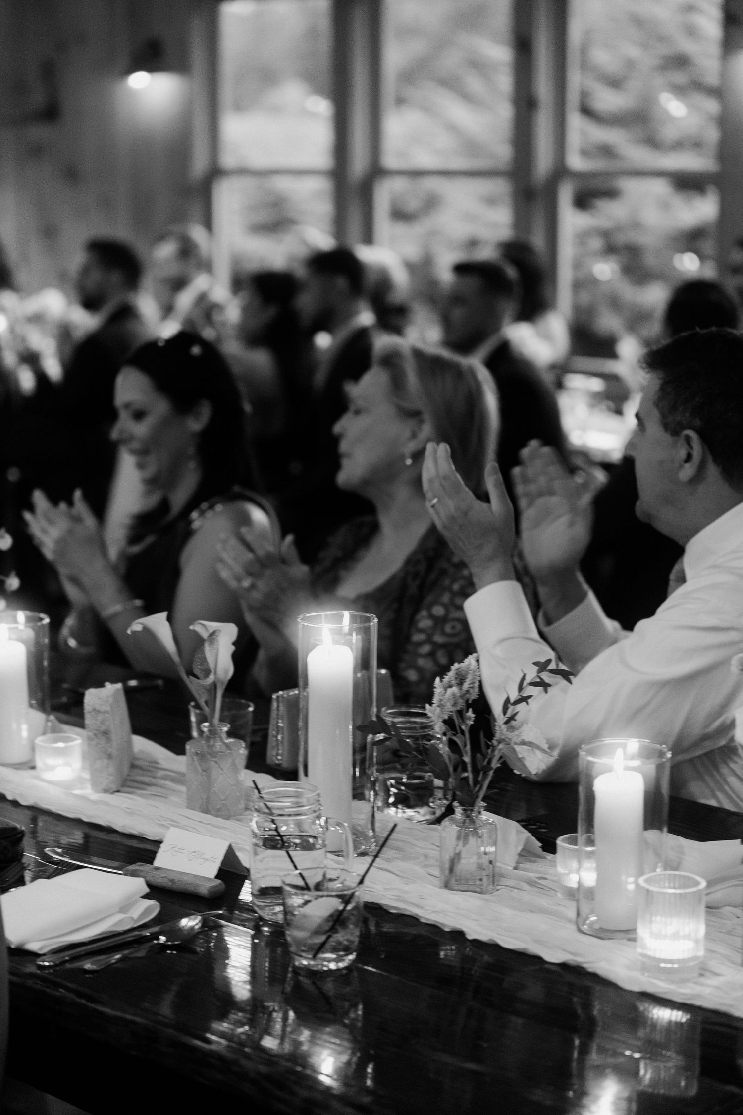 Black and white photo of wedding guests applauding at candlelit reception dinner table.