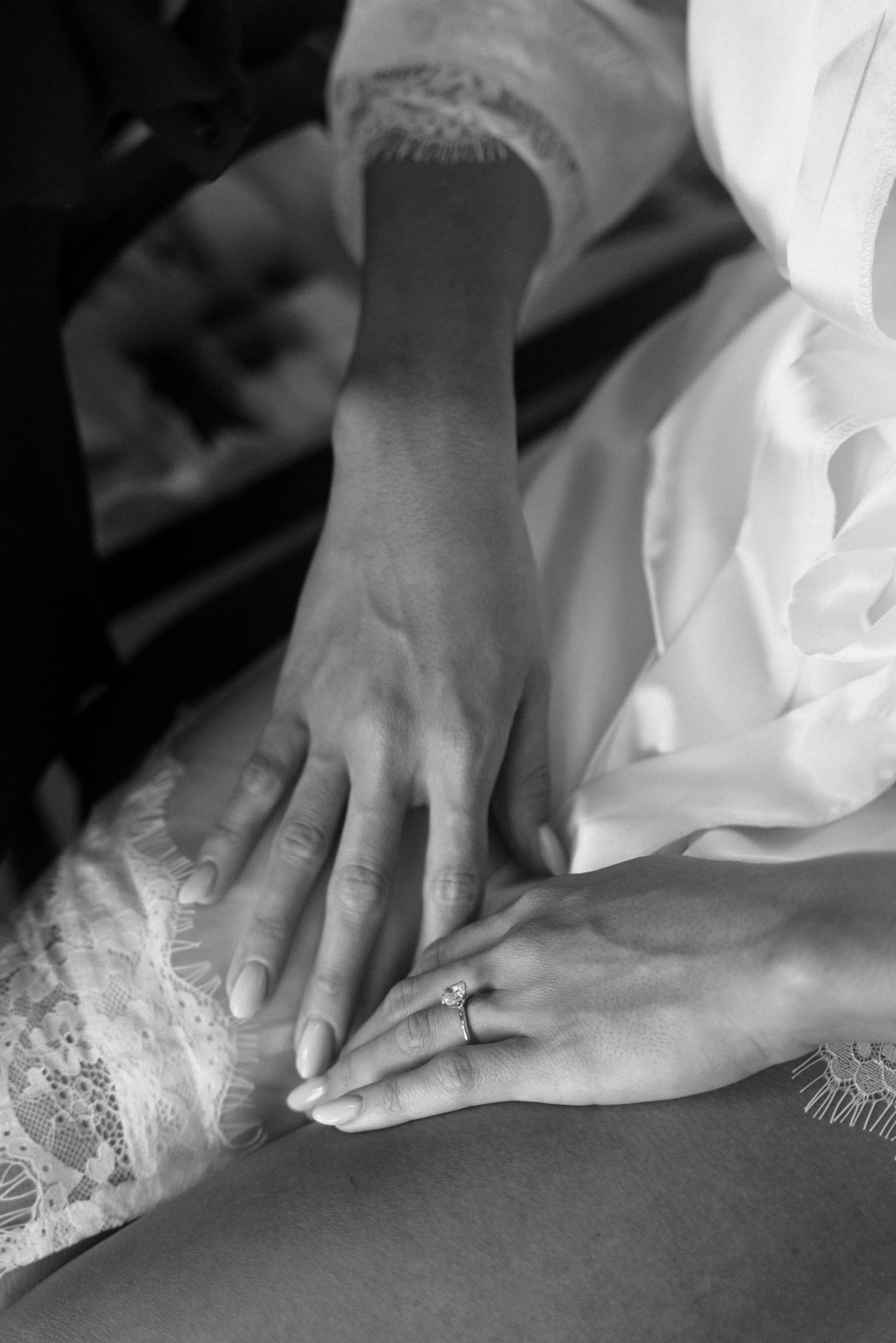 Black and white photo of hands with engagement ring resting on lace wedding dress fabric.