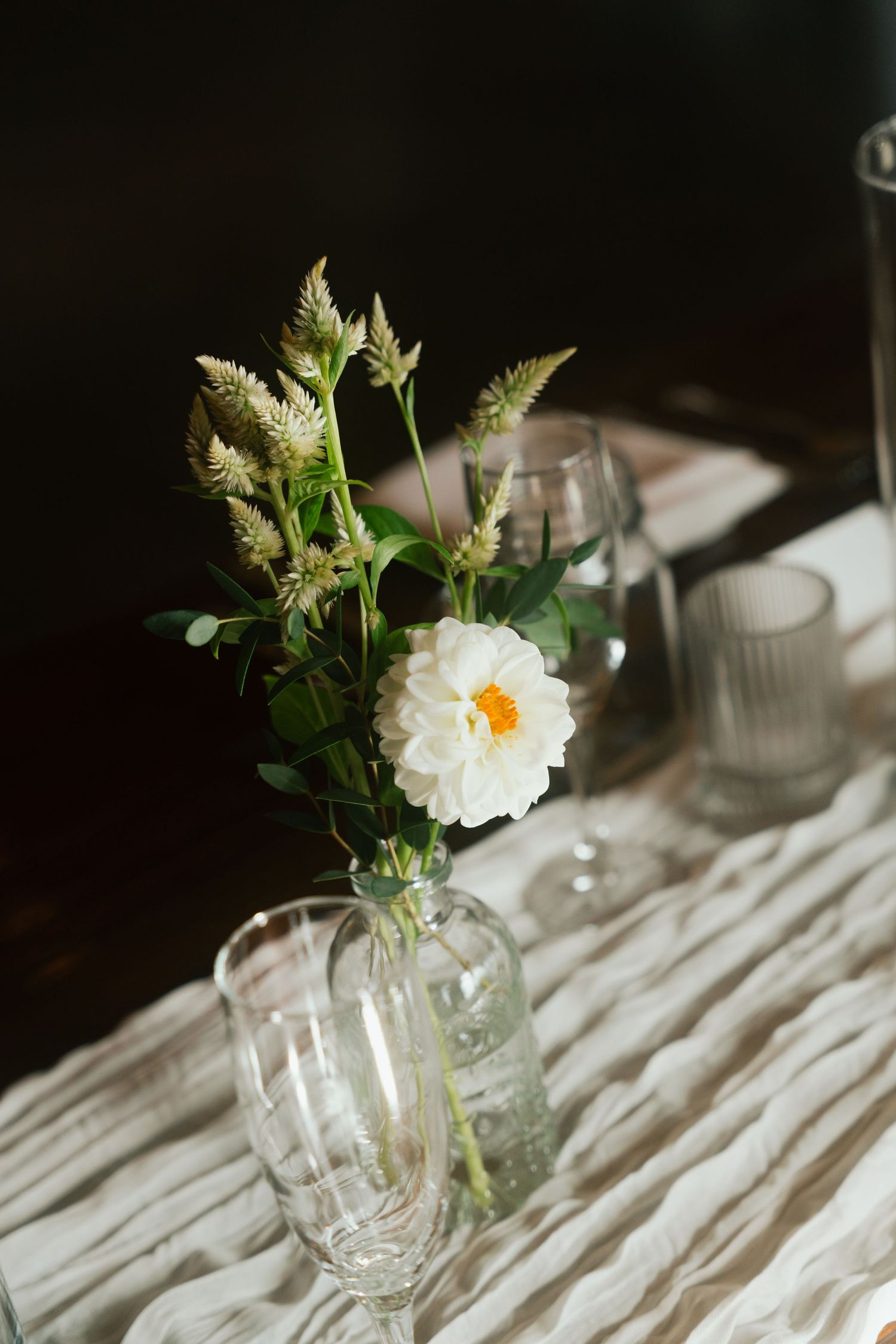 White peony and wildflowers in glass vase on linen table runner with glassware at wedding reception.