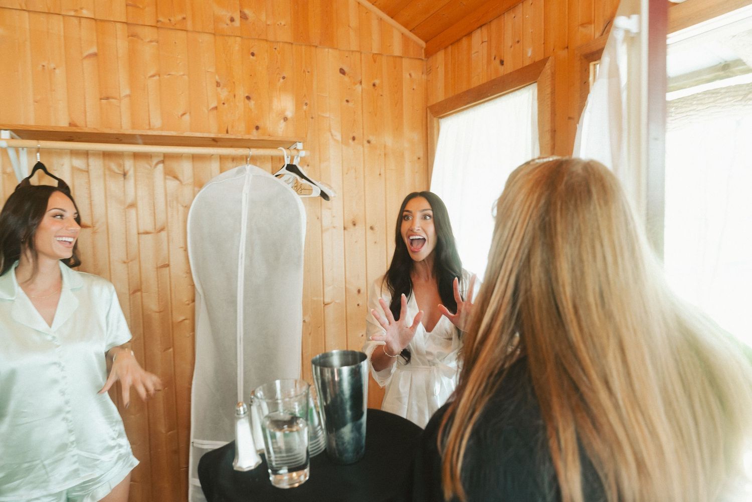 Two women in satin robes laughing together in a wooden cabin room with a wedding dress hanging nearby.