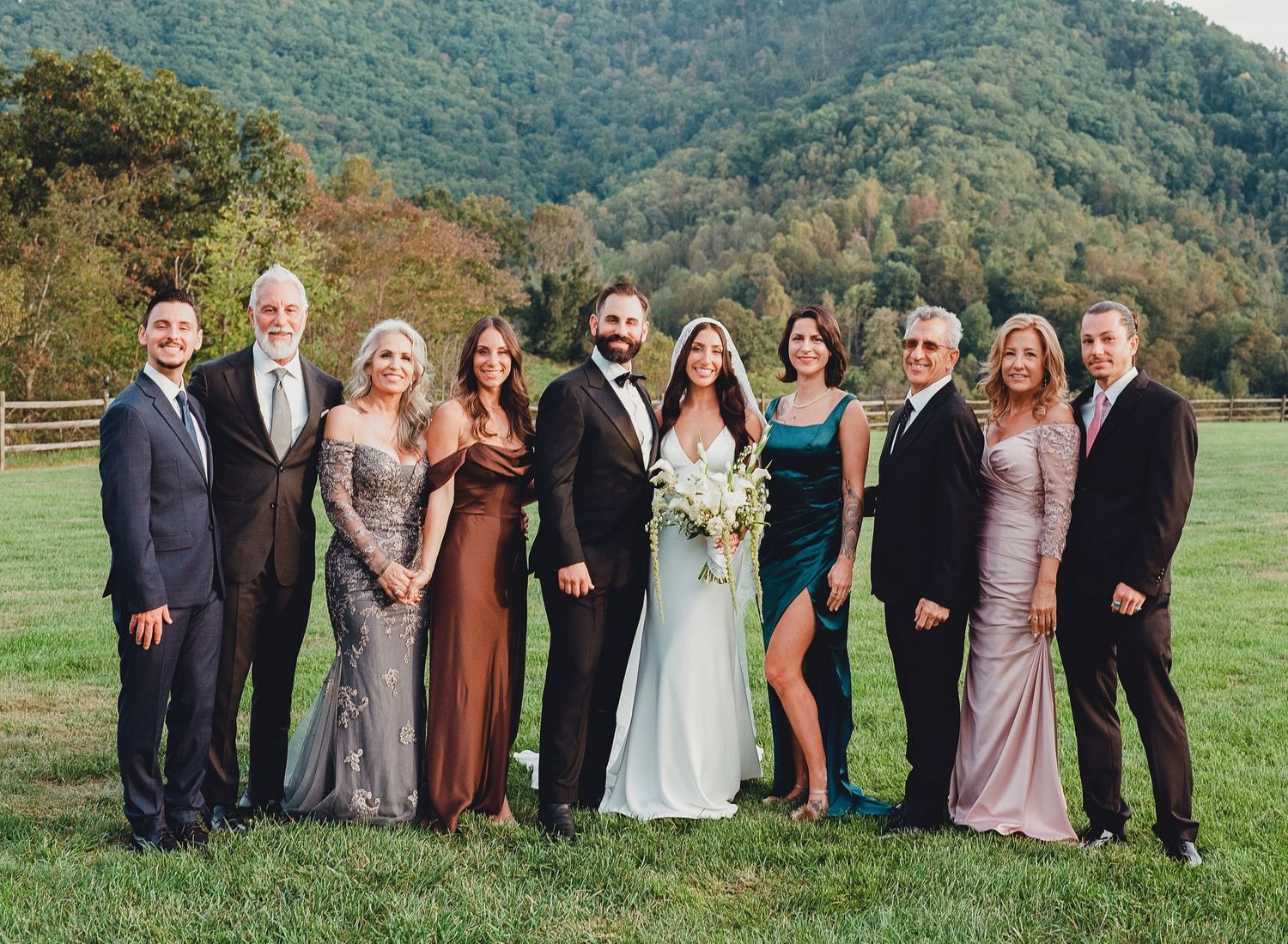 Wedding party poses outdoors with mountain backdrop, bride in white gown with bouquet, guests in formal attire.