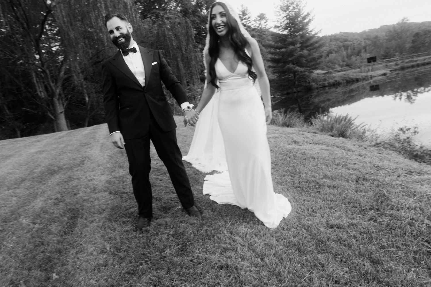 Black and white photo of newlyweds holding hands near a lake on their wedding day.