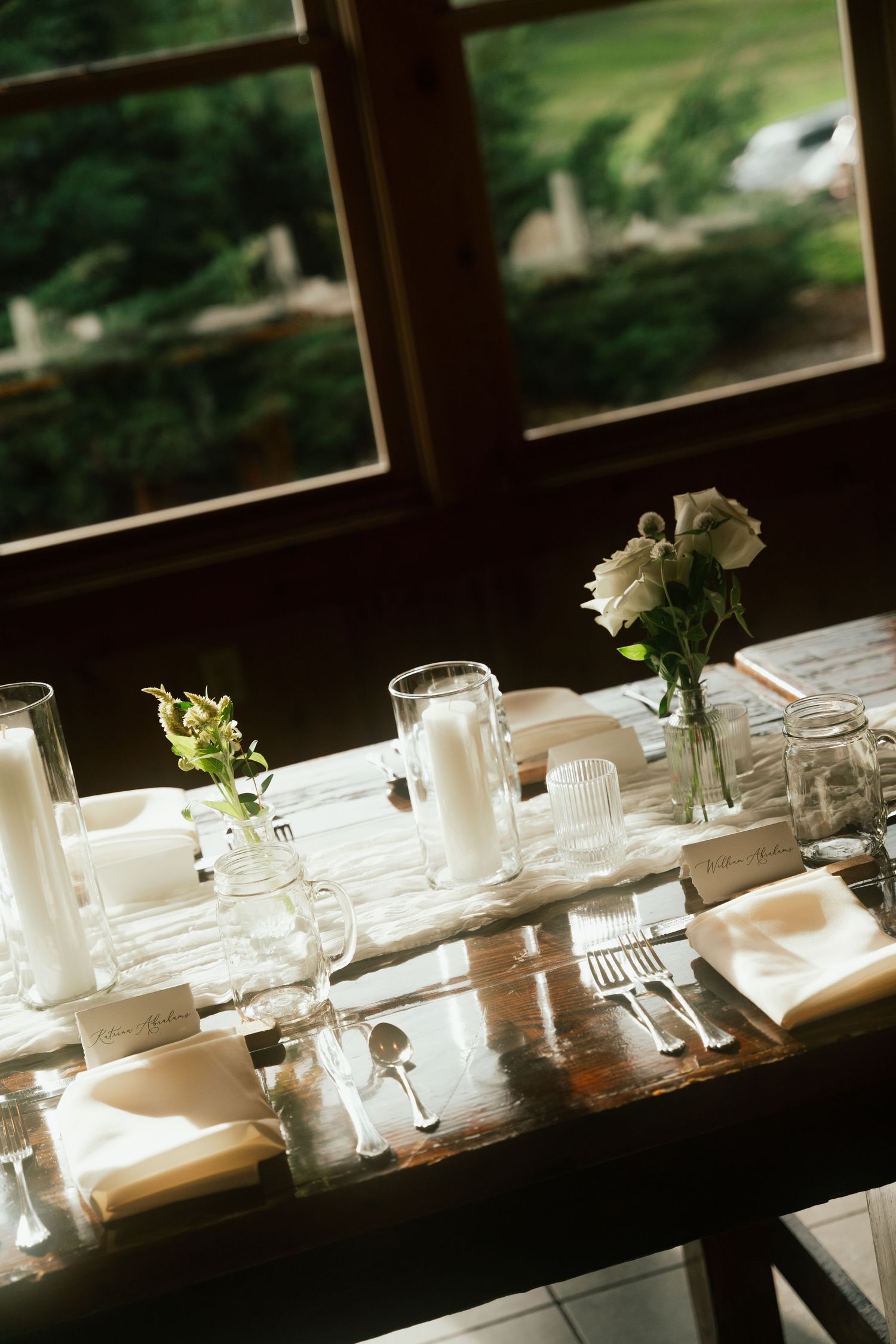 Elegant rustic wedding reception table set with white florals, candles, and glassware by a window.