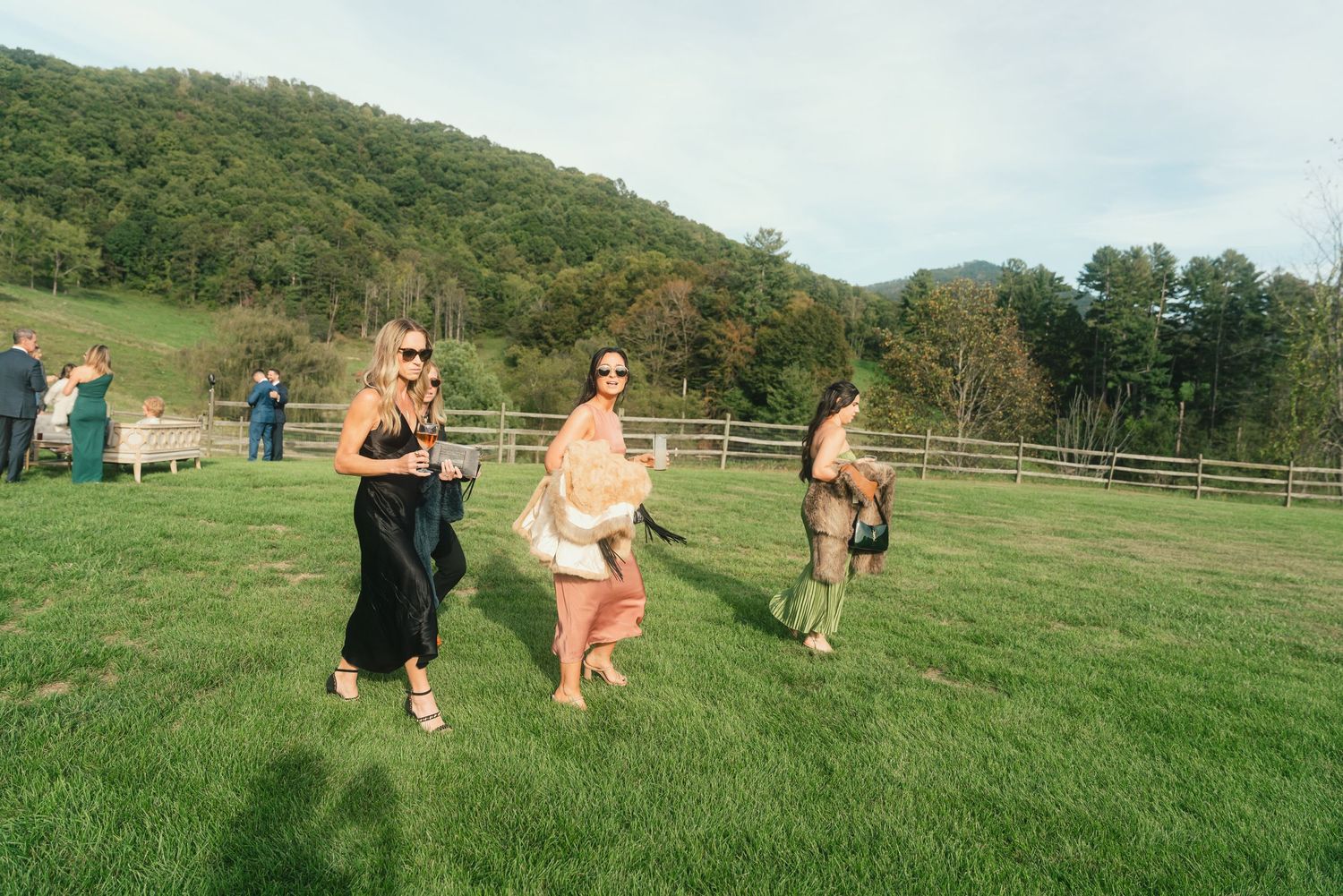 Elegantly dressed guests walk across a green lawn with rolling mountain hills in the background at an outdoor event.
