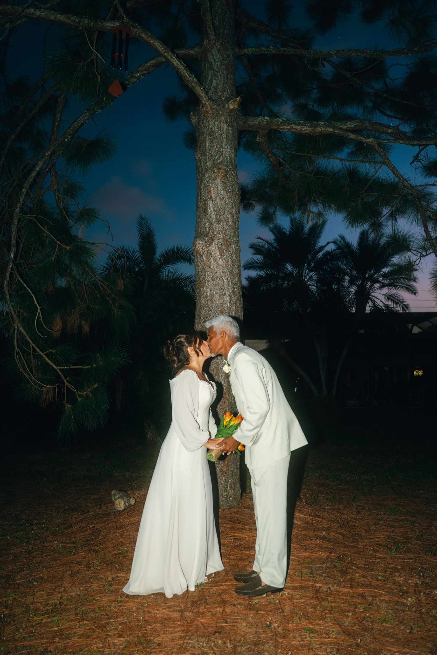 A couple in white formal wear sharing an intimate moment in a wooded setting at twilight.