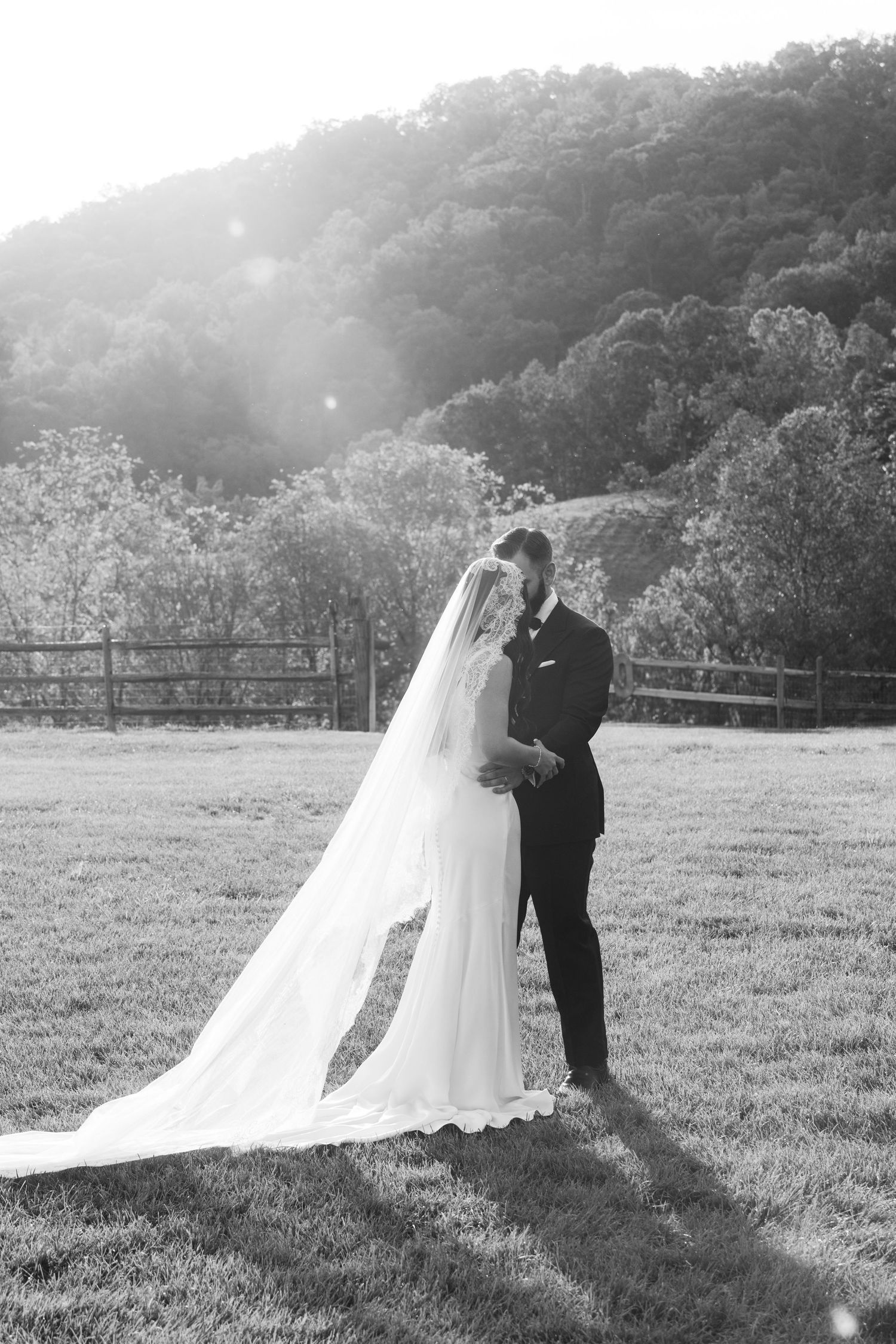 Wedding couple embracing in a sunlit meadow with mountains in the background.