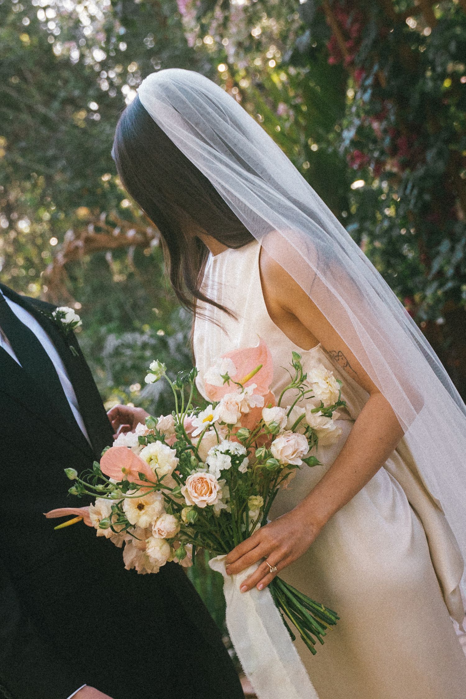 A bride in white dress holds a delicate bouquet of pink and white flowers against a sunlit garden backdrop.