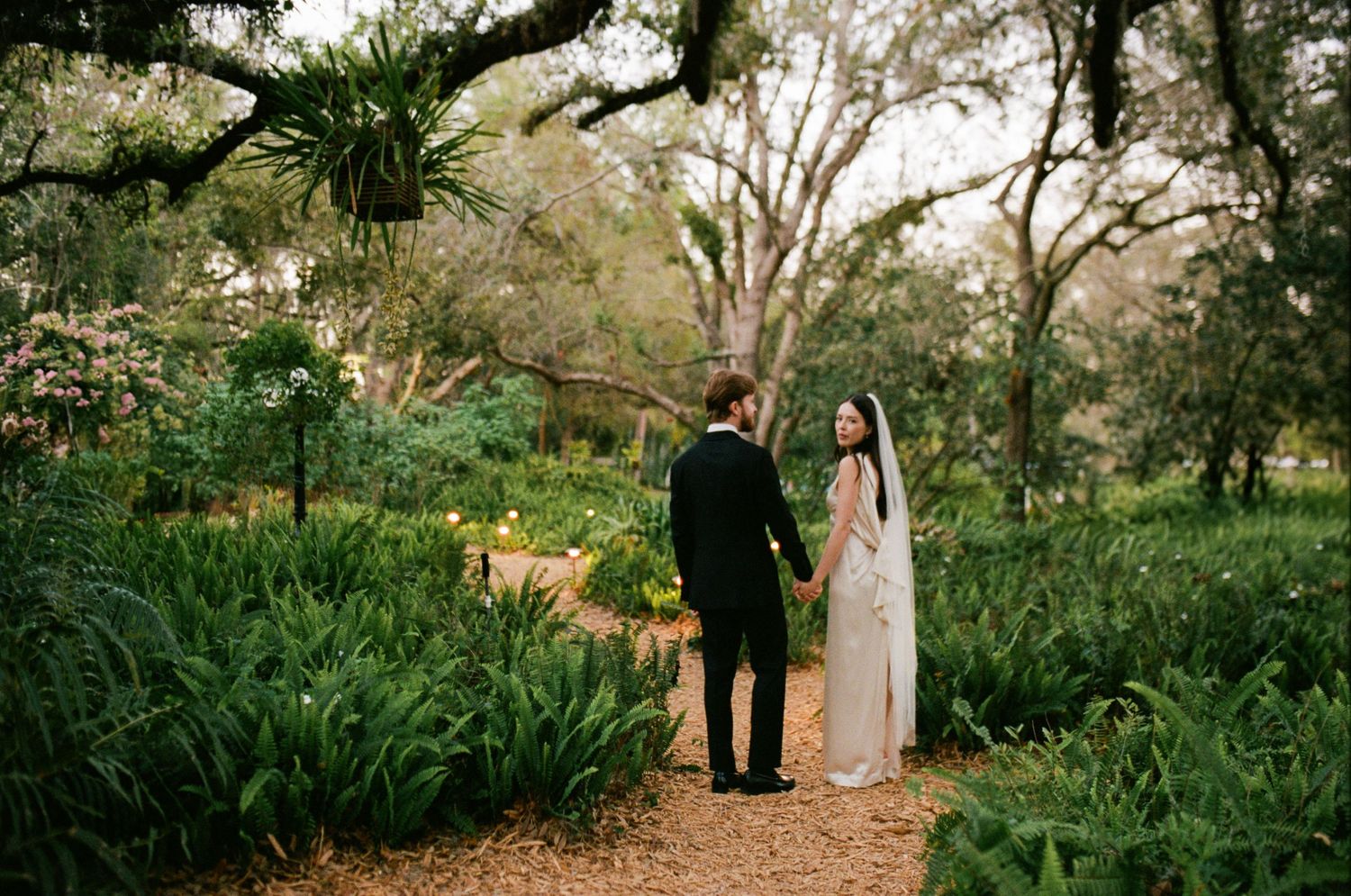 A couple walks down a garden path surrounded by lush greenery and trees.