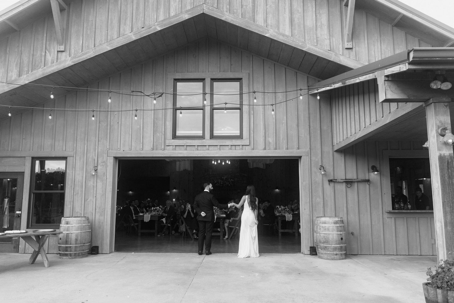 Black and white photo of a bride and groom standing outside a rustic barn wedding venue with string lights.