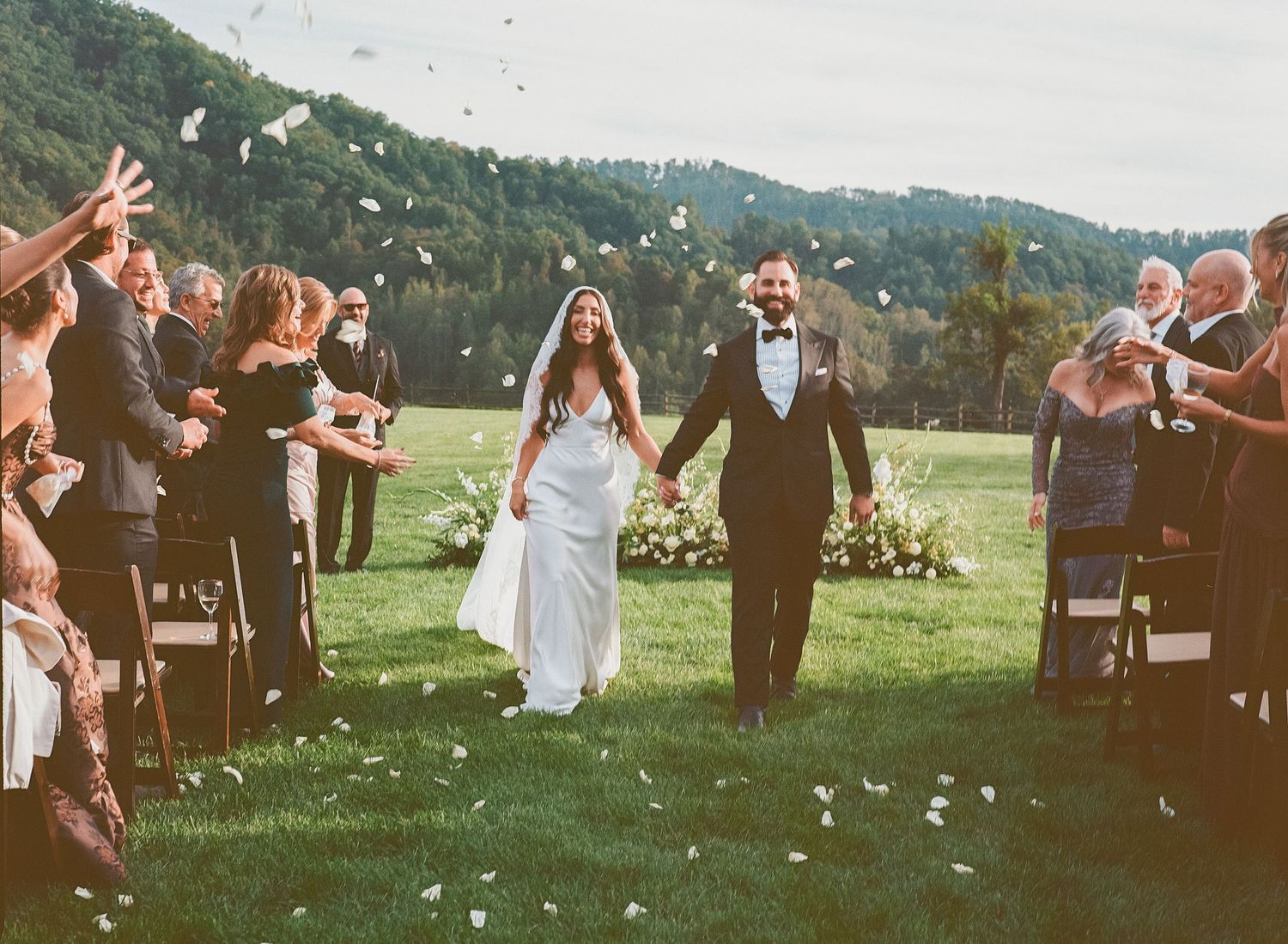 Bride and groom walk down aisle as guests toss white flower petals at outdoor wedding ceremony in scenic mountain setting.