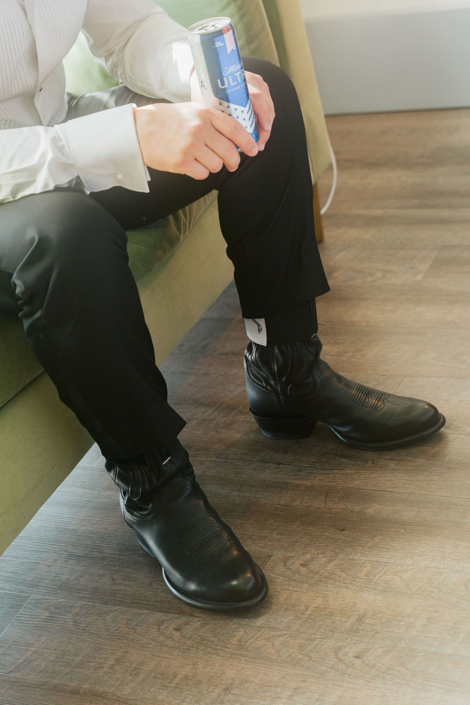 Man in black pants and cowboy boots sitting on green couch holding a Red Bull energy drink can.