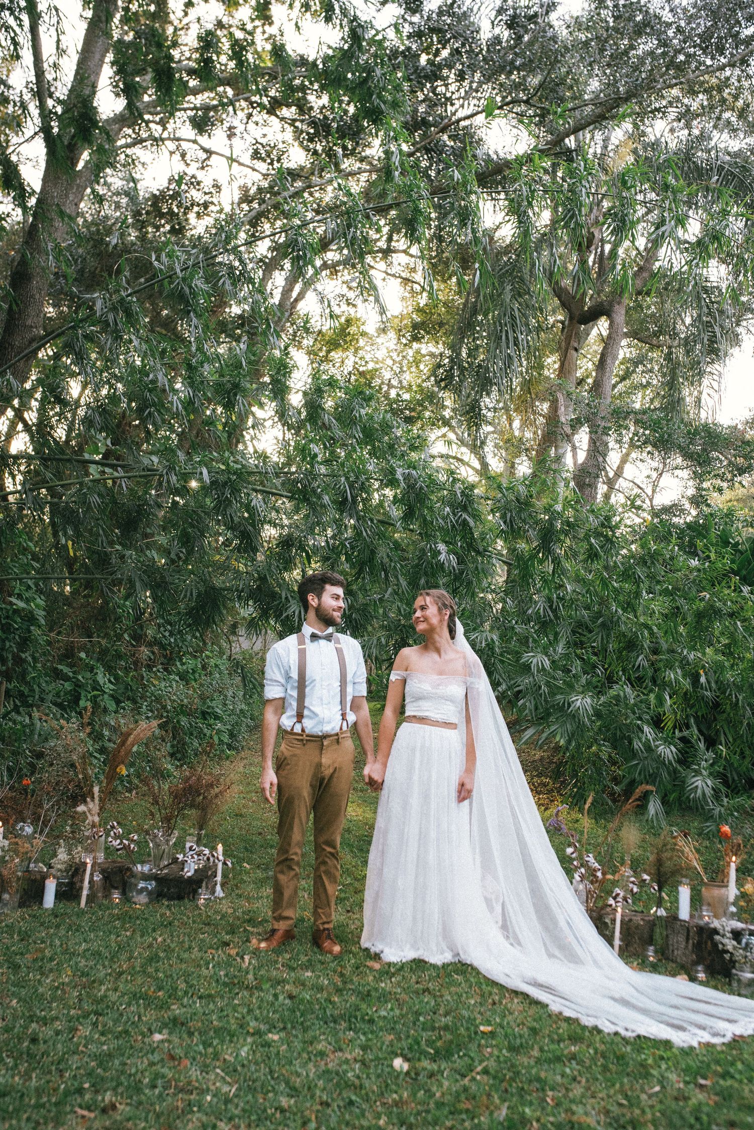 Wedding couple standing under trees with decorative glass bottles and candles in background.