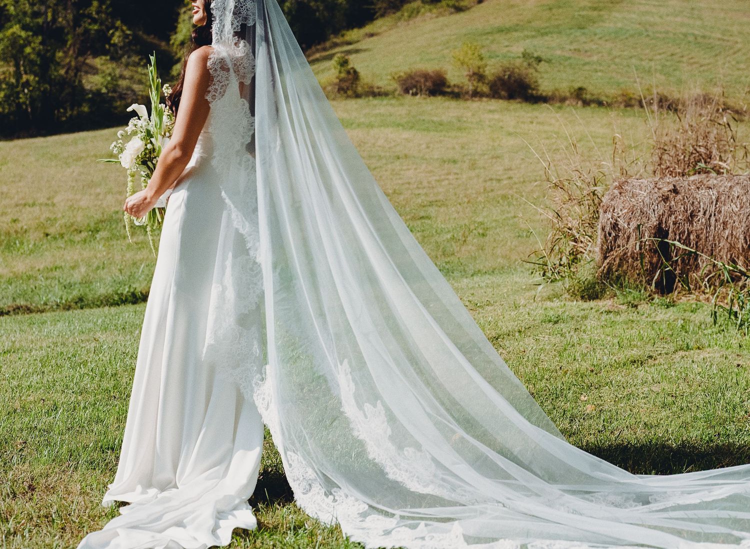 Bride in elegant white wedding gown with long flowing veil standing on green grass holding a bouquet.