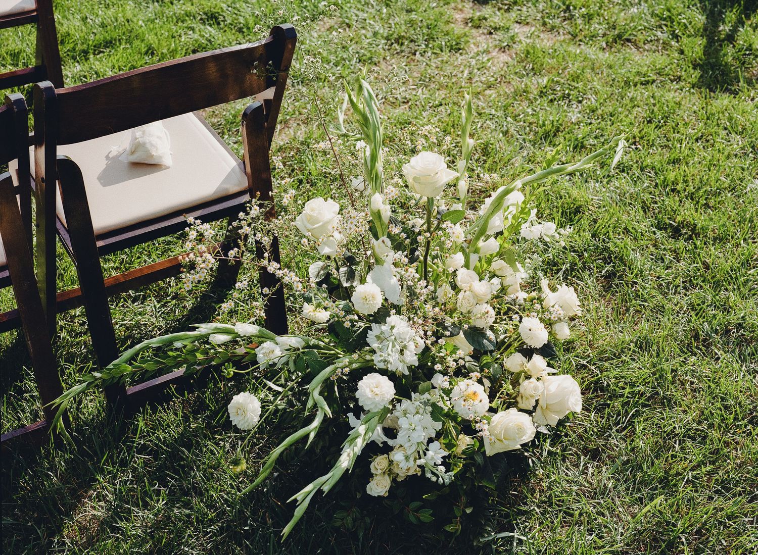 Elegant white floral arrangement with roses and greenery placed beside wooden chair at outdoor wedding ceremony.