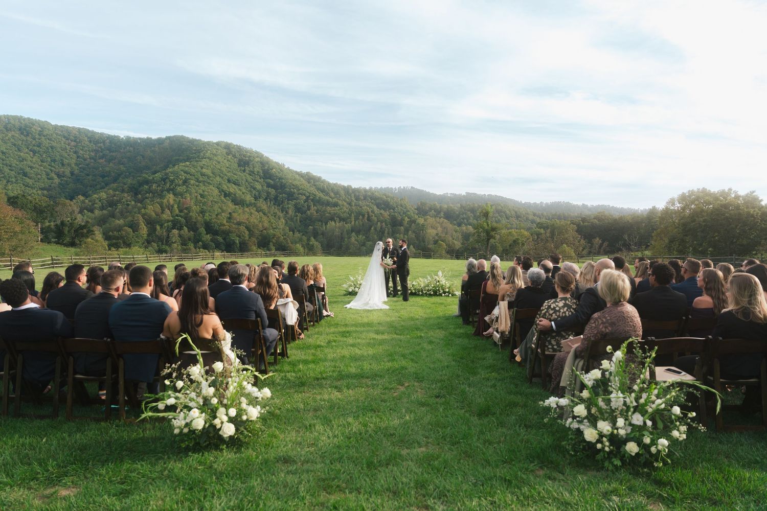 Wedding guests seated in rows on a hilltop with mountain views and floral decorations.