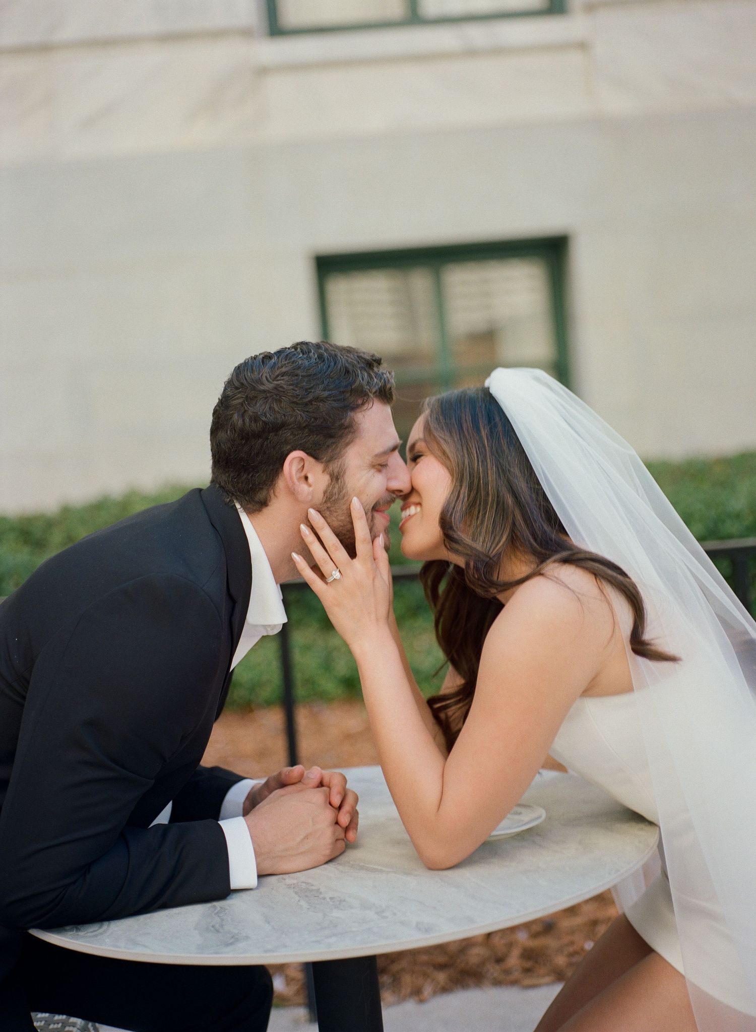 Bride and groom share a romantic almost-kiss moment at a marble table outdoors on wedding day.