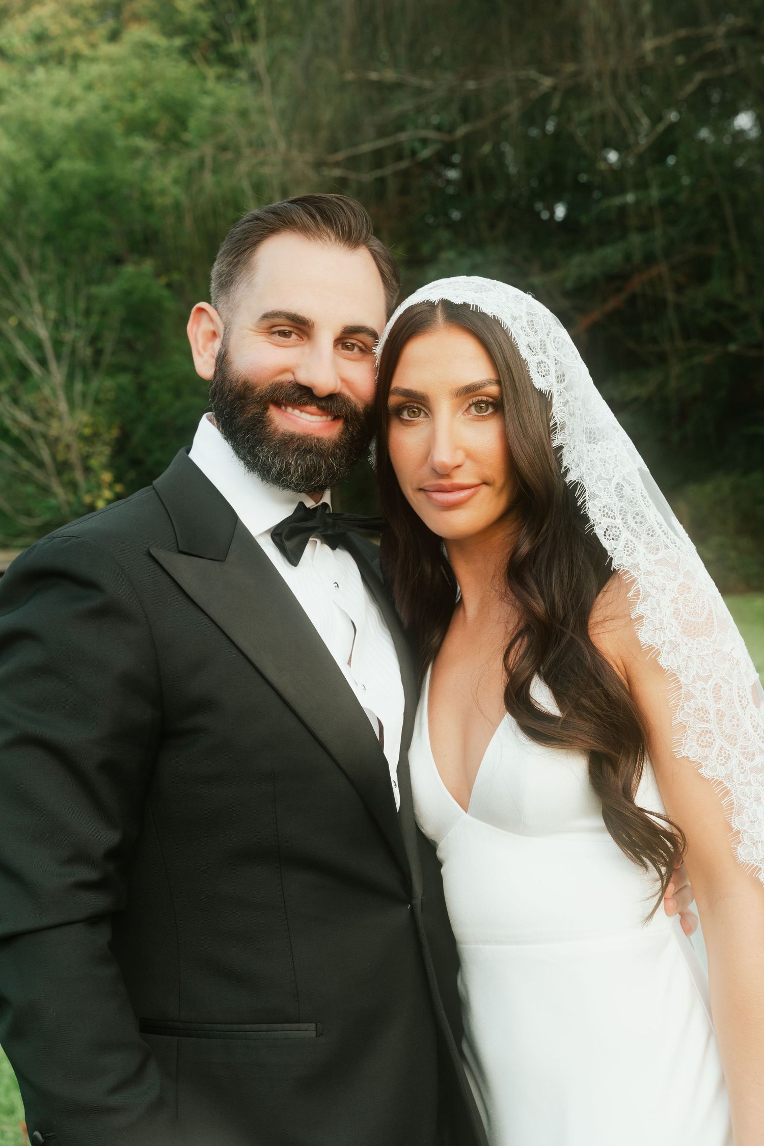 A smiling bearded groom in a black tuxedo poses with his bride in a white gown and lace veil outdoors.
