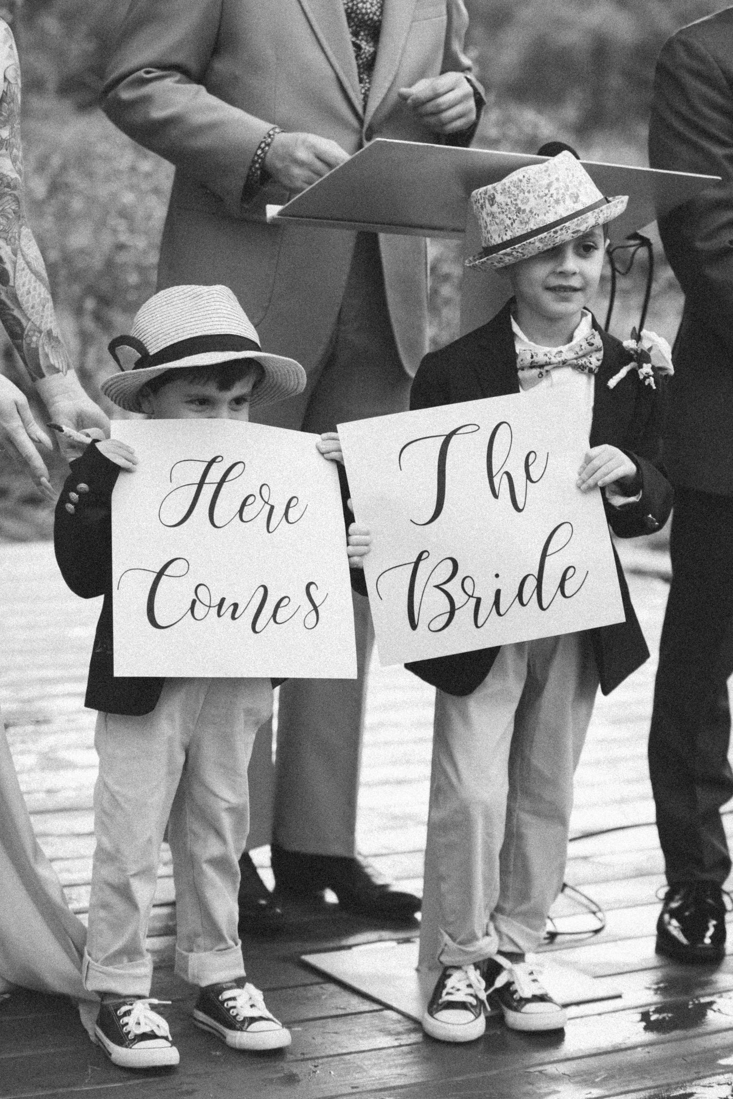 Two young children in formal attire hold signs reading 'Here Comes The Bride' at a wedding ceremony.