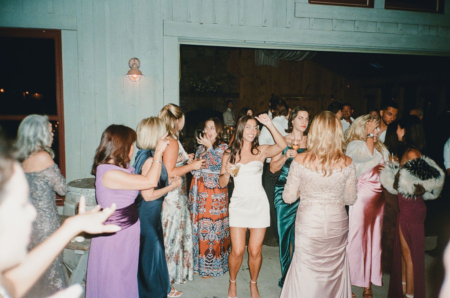 Guests in elegant dresses dance together joyfully at an indoor wedding reception celebration.