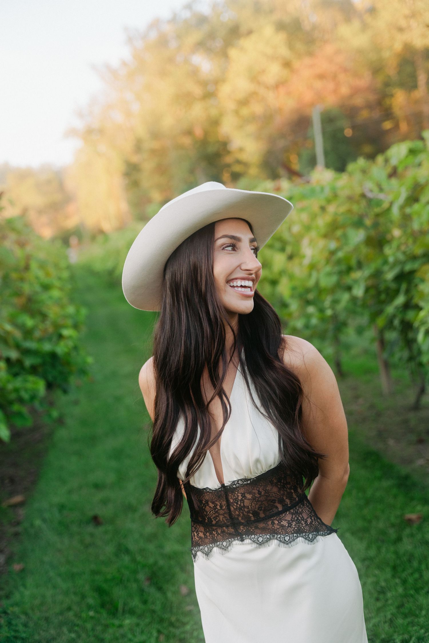 Smiling woman in white cowboy hat and lace dress standing in lush green vineyard rows on a sunny day.