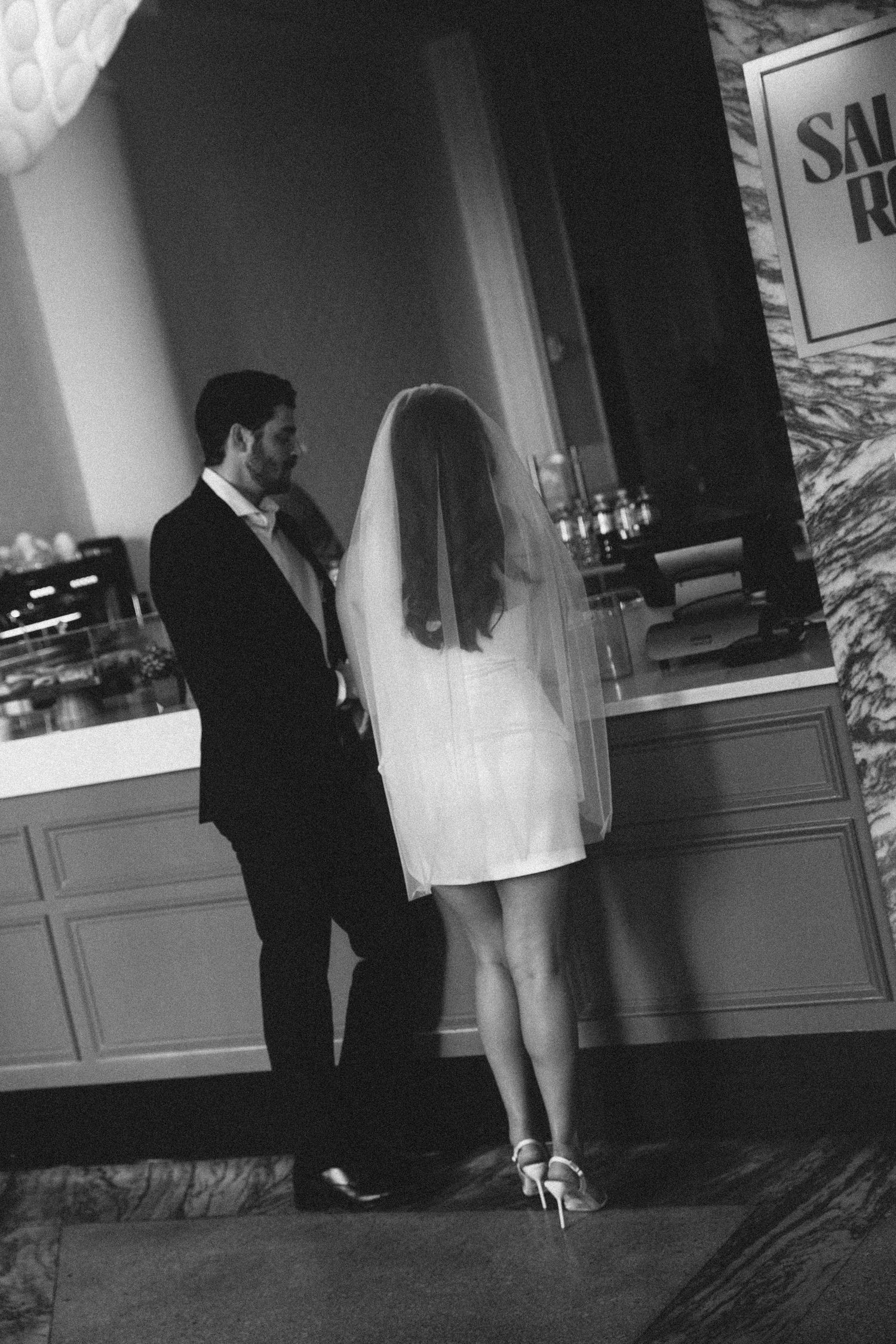 Black and white photo of bride in mini dress with veil and groom standing together at a bar.