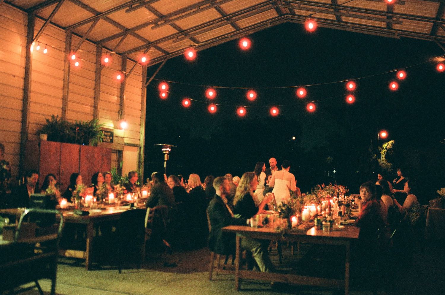 String lights illuminate an outdoor evening wedding reception under a wooden pavilion.