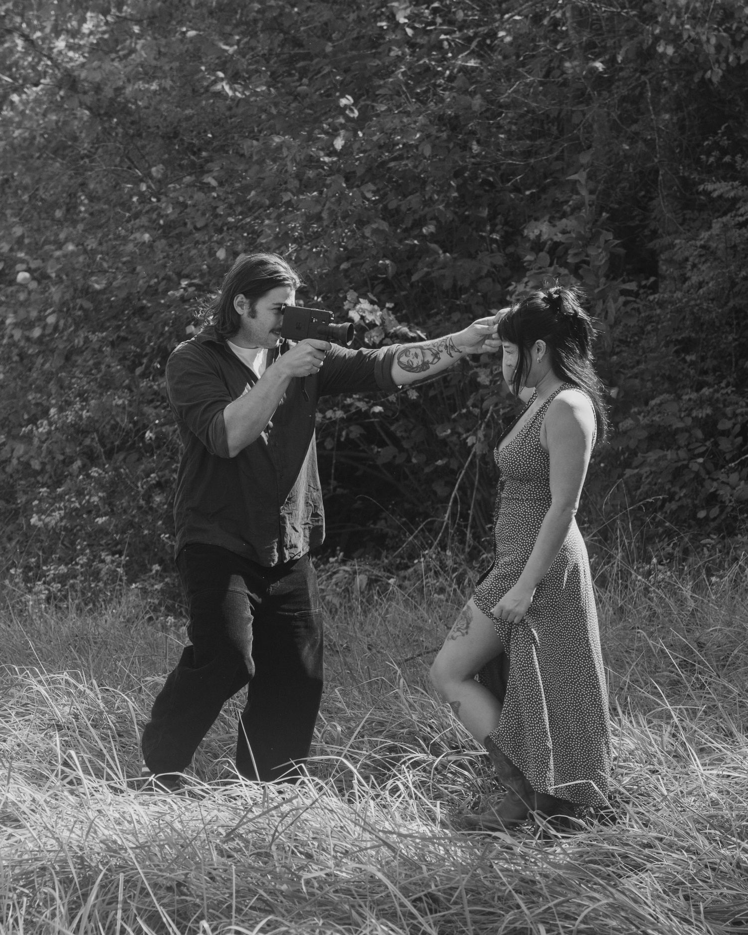 Black and white photo of a man pointing a camera at a woman in a field with trees in the background.