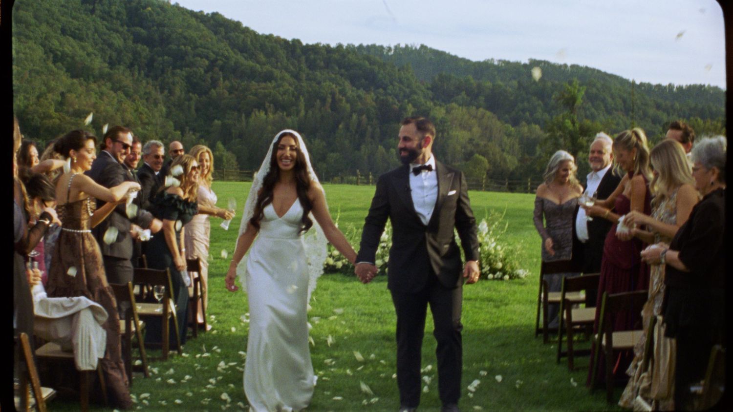 A wedding party celebrates outdoors in a lush green meadow surrounded by forest-covered hills in the background.