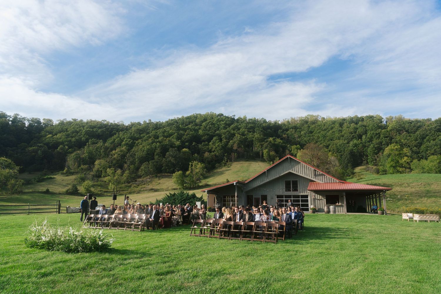 Outdoor wedding ceremony setup with wooden chairs on a green hillside near a rustic building.