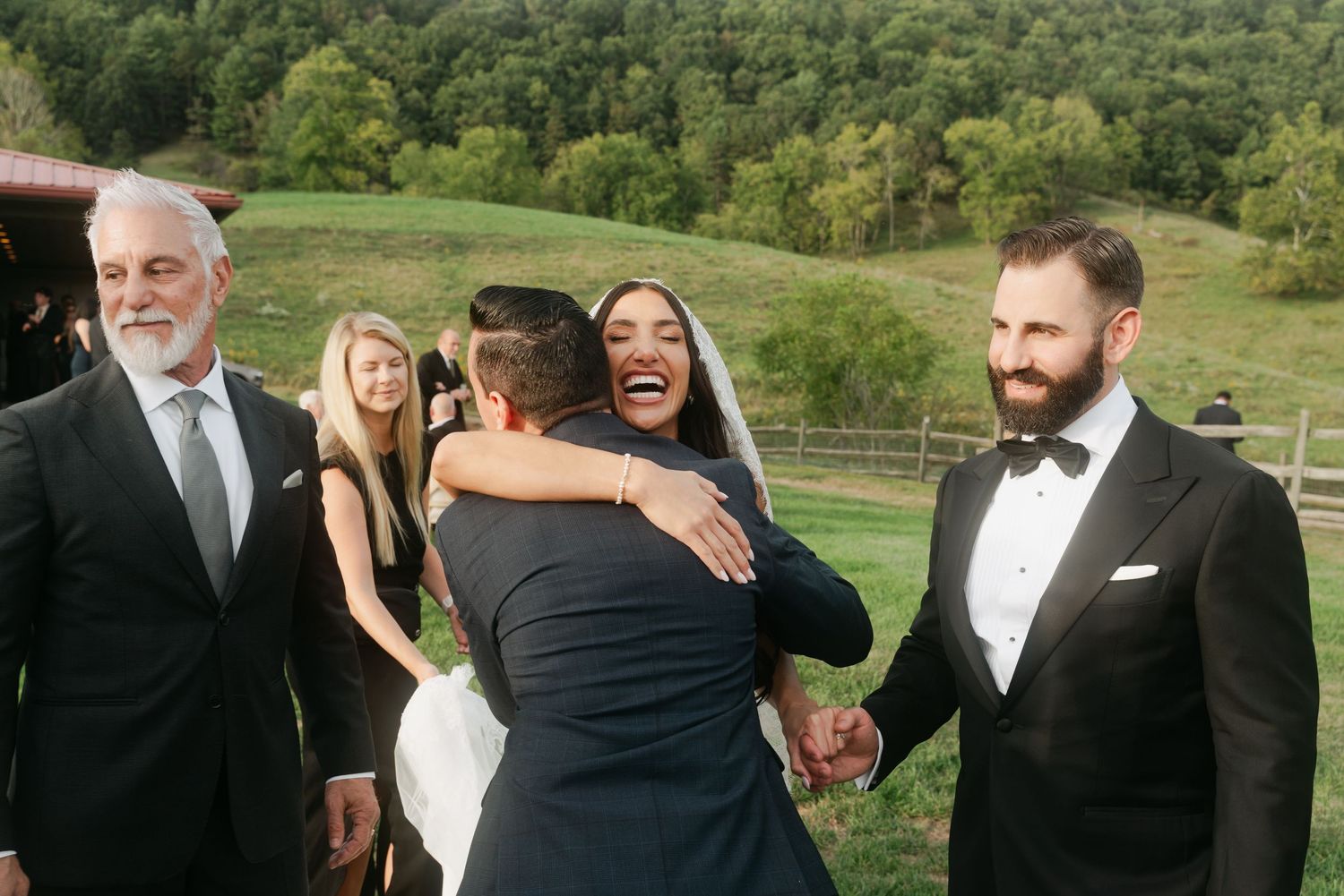 Wedding guests embracing and shaking hands outdoors, with lush green hills in the background at a countryside ceremony.
