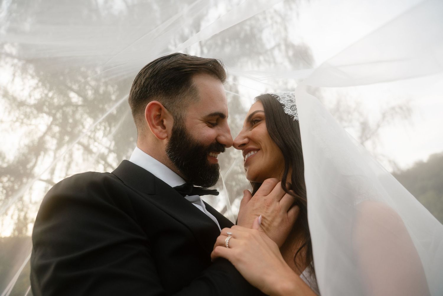 A bride and groom share a romantic moment under a flowing veil, nearly kissing outdoors on their wedding day.