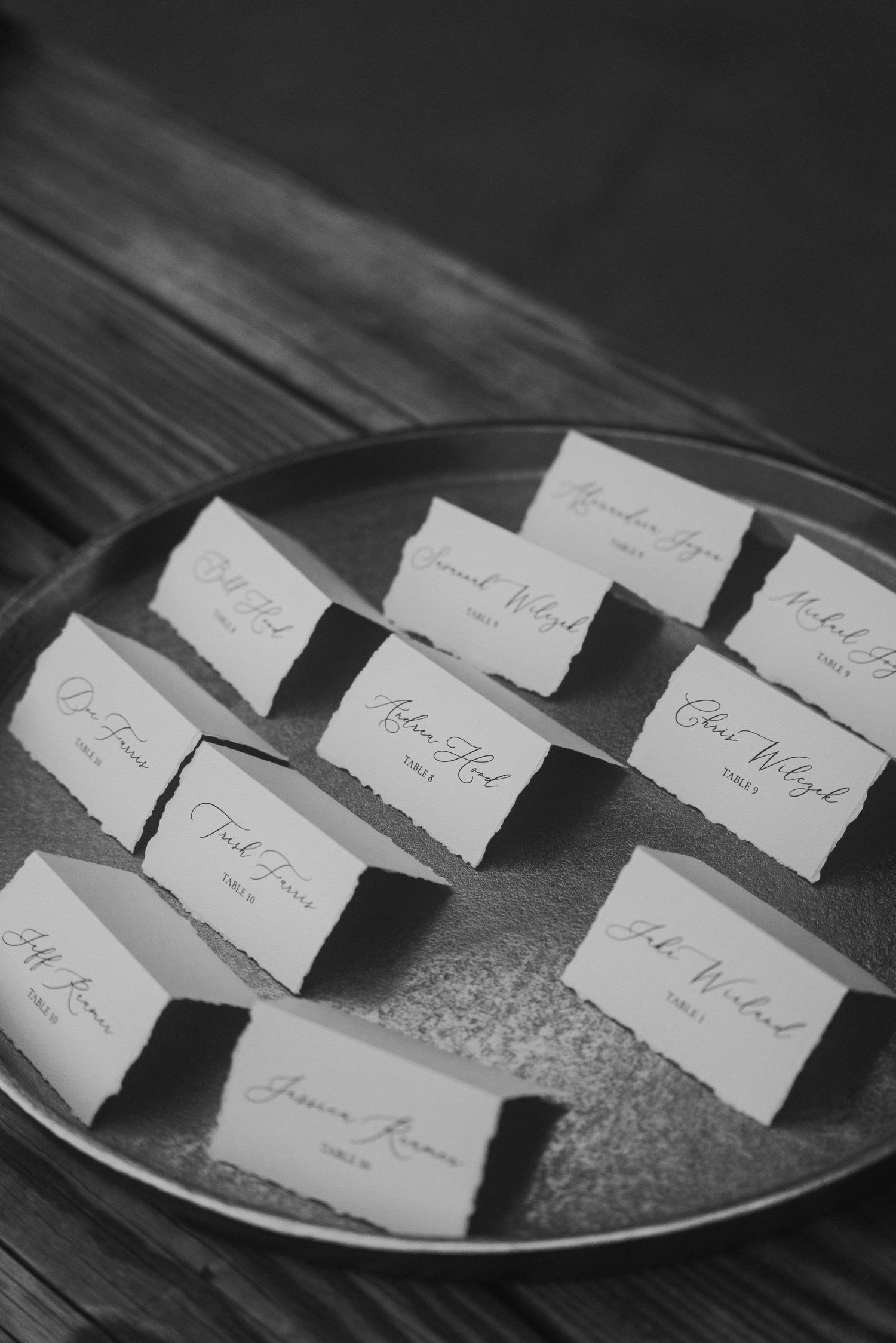 Black and white photo of elegant handwritten wedding escort cards displayed on a round metal tray on a wooden table.