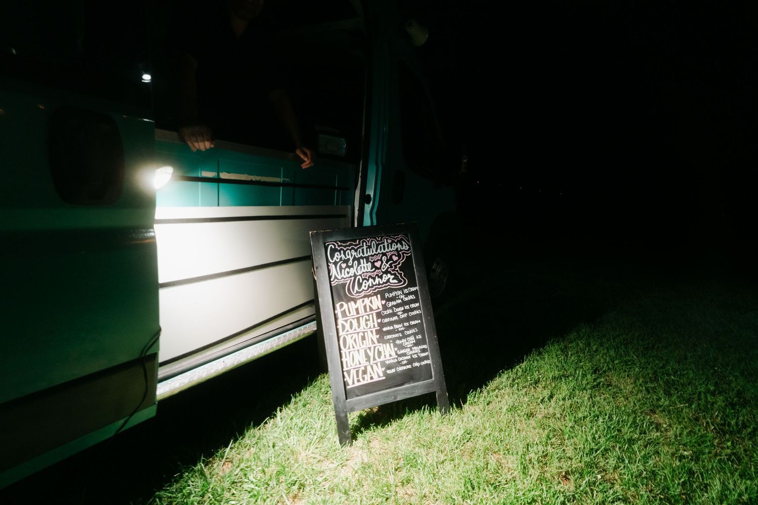 A chalkboard sign glows beside a food truck at an outdoor nighttime wedding reception on green grass.