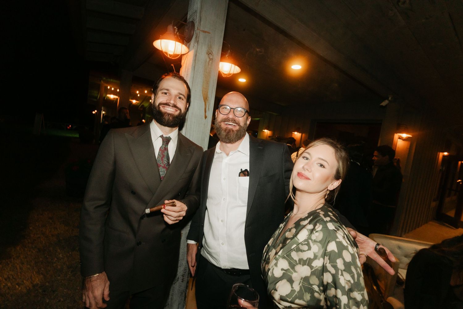 Three wedding guests pose together outdoors at night near warm glowing lights at reception.