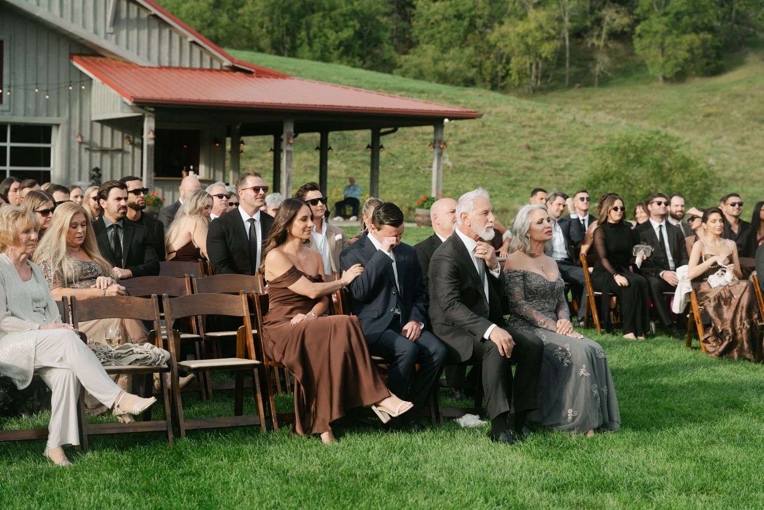 Guests seated outdoors on green lawn chairs at a rustic barn wedding ceremony on a sunny day.