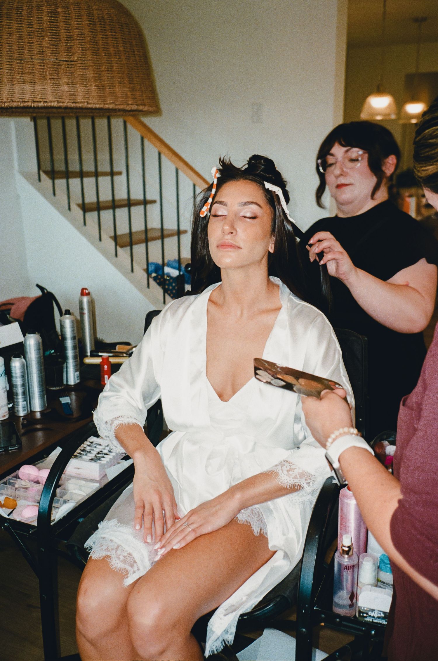 Bride in white lace robe gets hair and makeup done by stylists before her wedding day.
