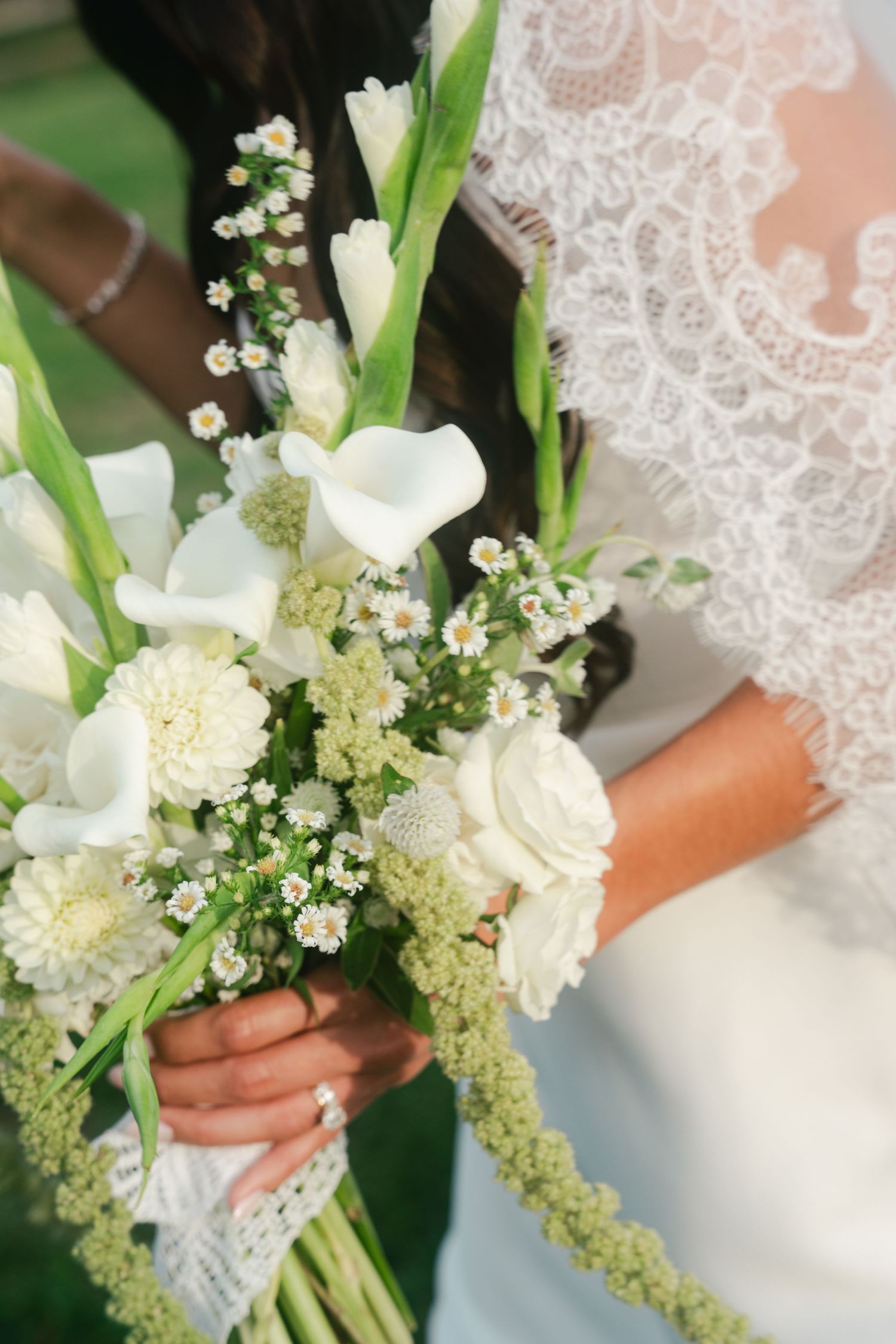 Bride in lace dress holding elegant white wedding bouquet with calla lilies, roses, and wildflowers.