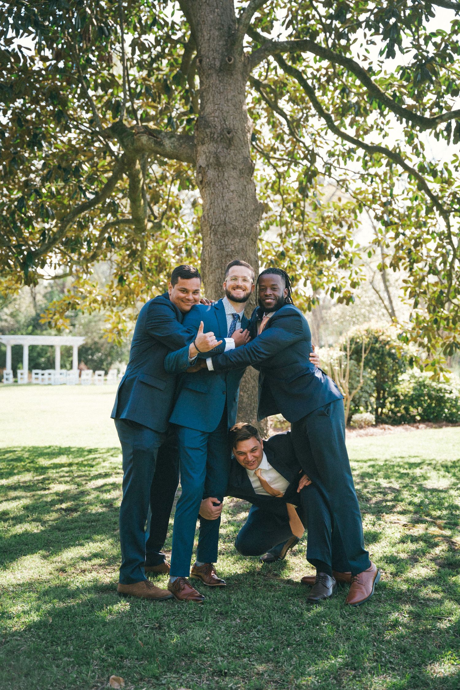 Groomsmen in blue suits playfully posing together under a tree in a garden setting.