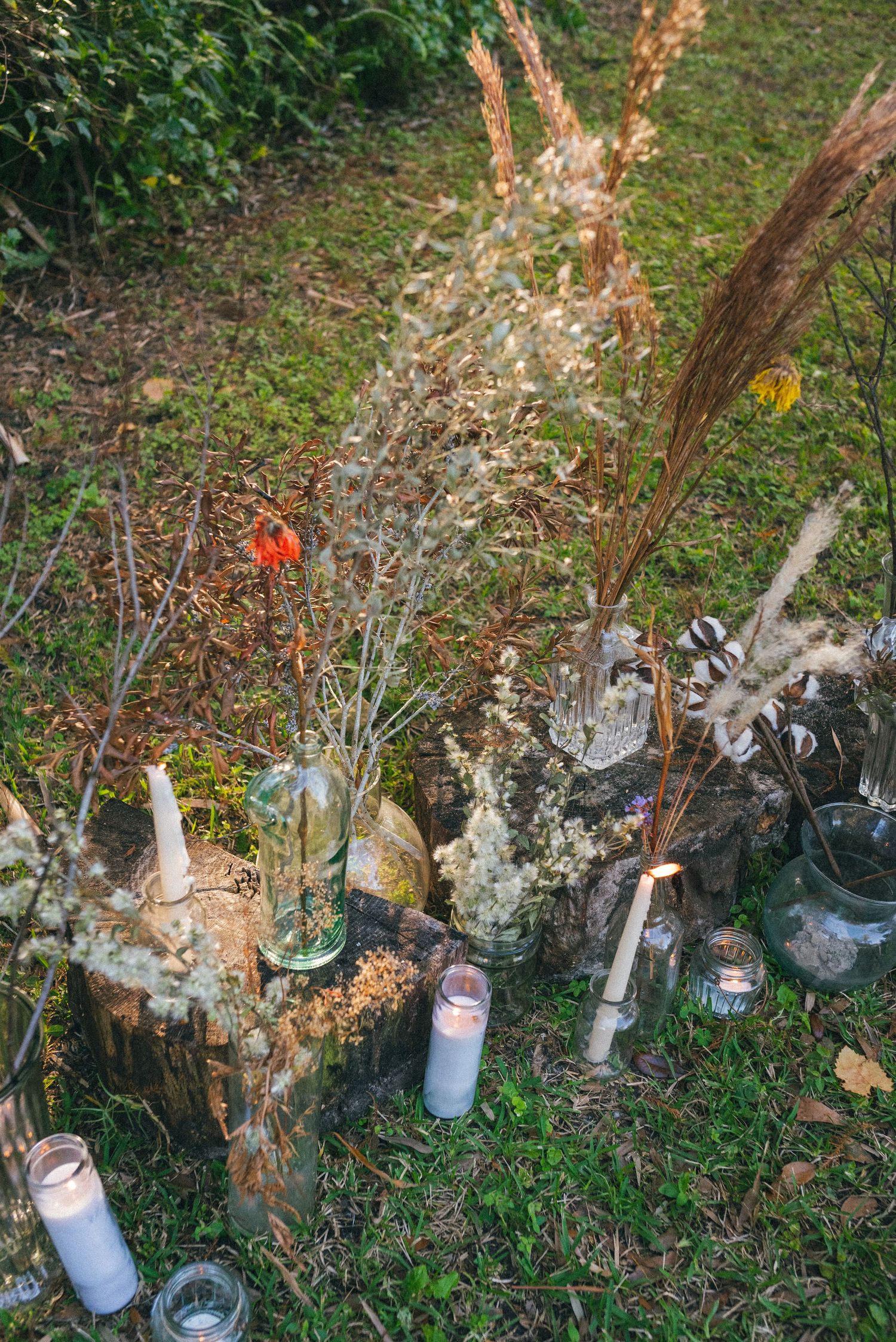 Outdoor wedding decor featuring vintage glass bottles, candles and dried flowers on grass.