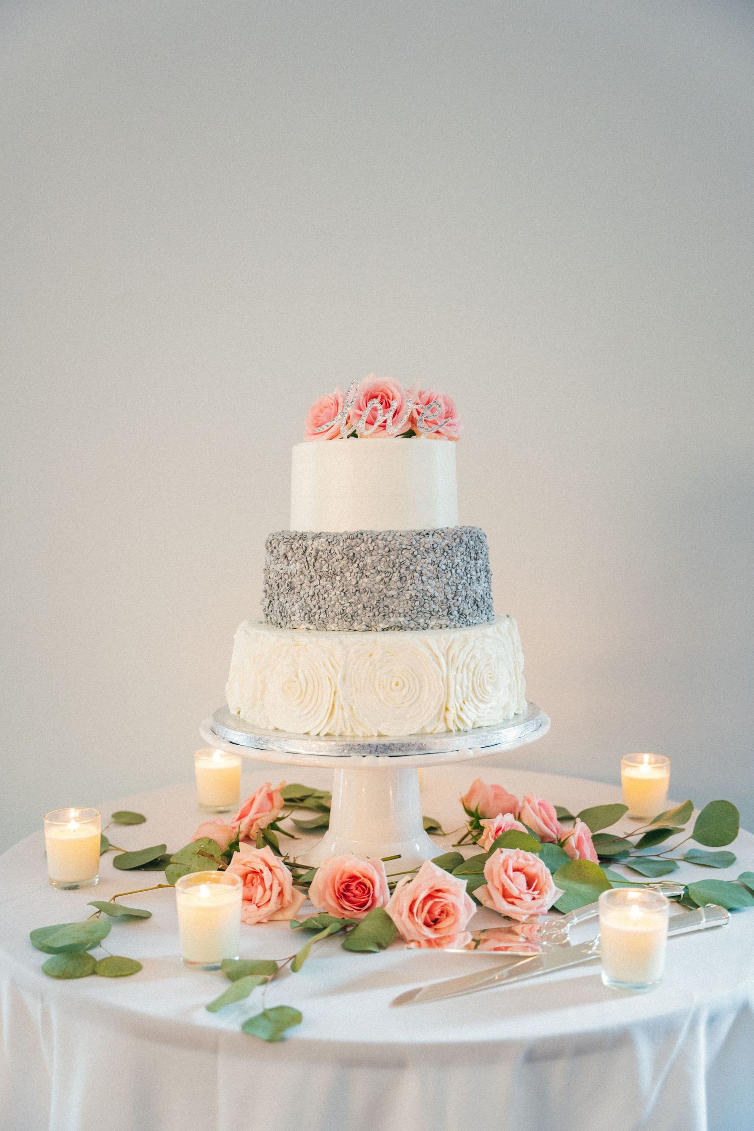 Elegant wedding cake display with pink roses and candles on white tablecloth.